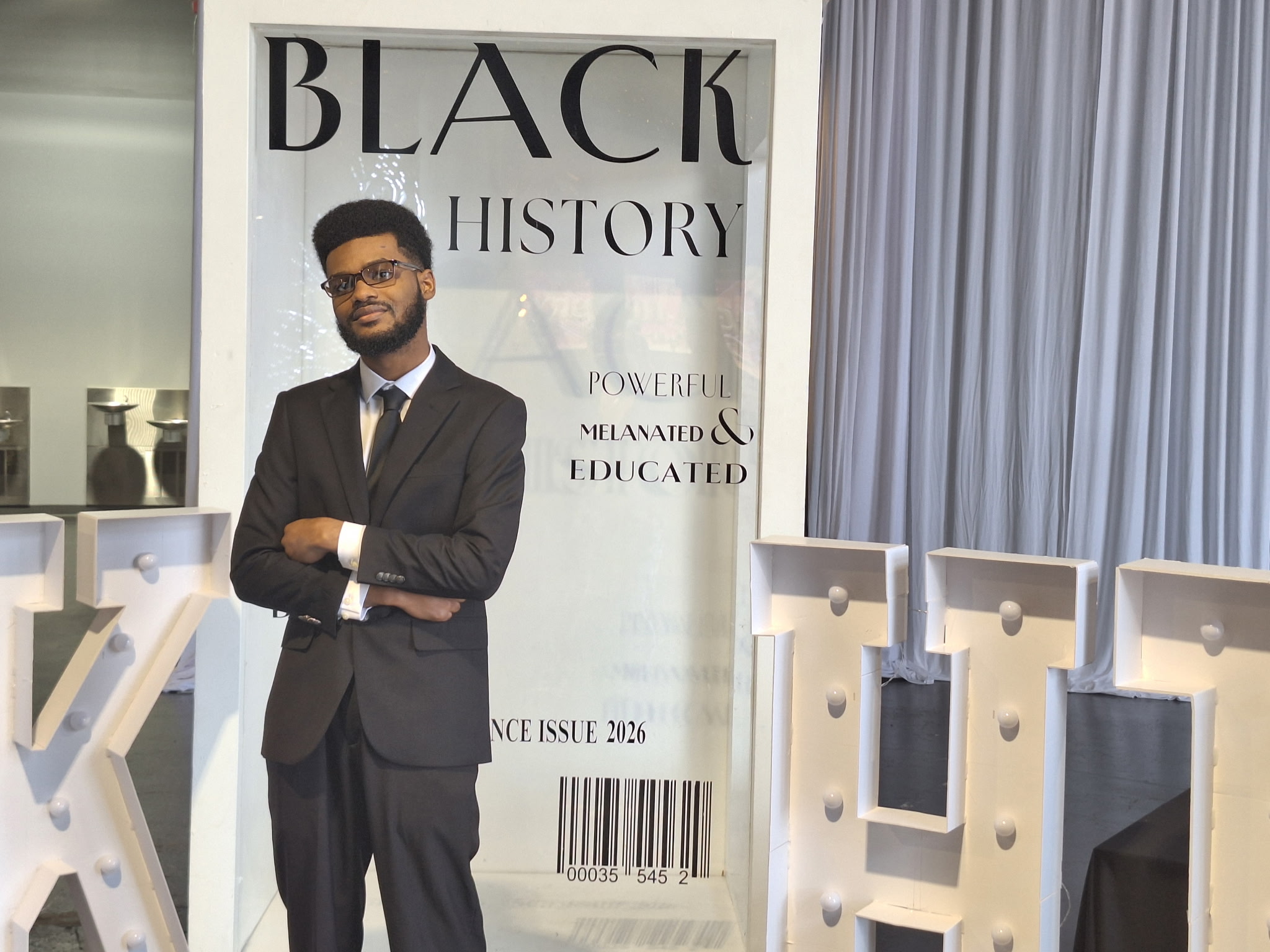 Young man in a suit standing in front of a poster about Black history, with blue curtains nearby.