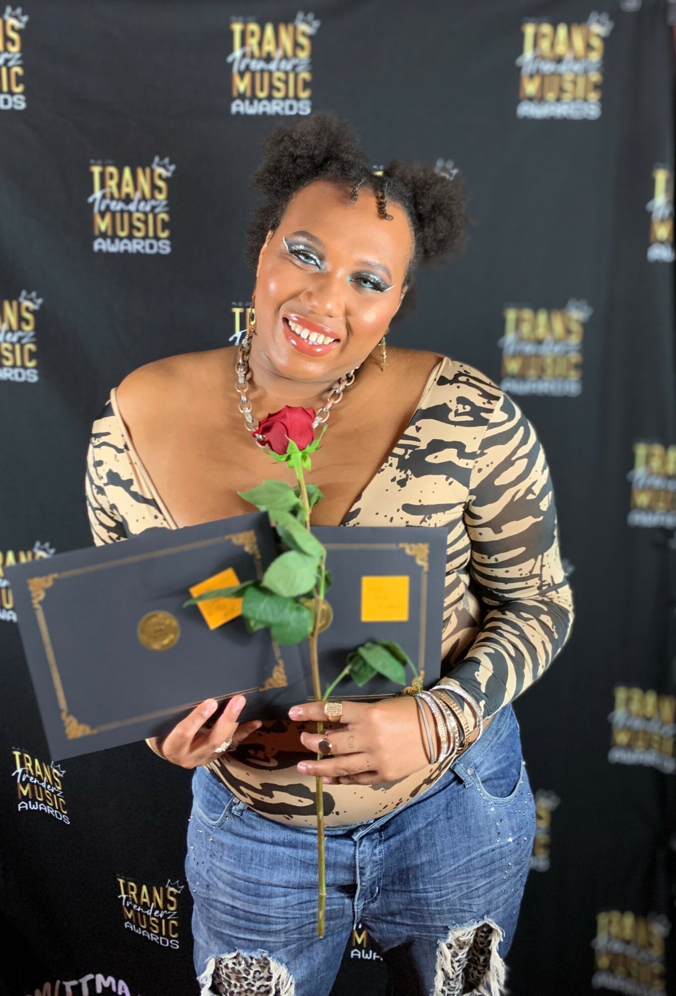 Woman smiling holding a rose and a certificate at an awards event, background with 'Trans Music Awards' logos.