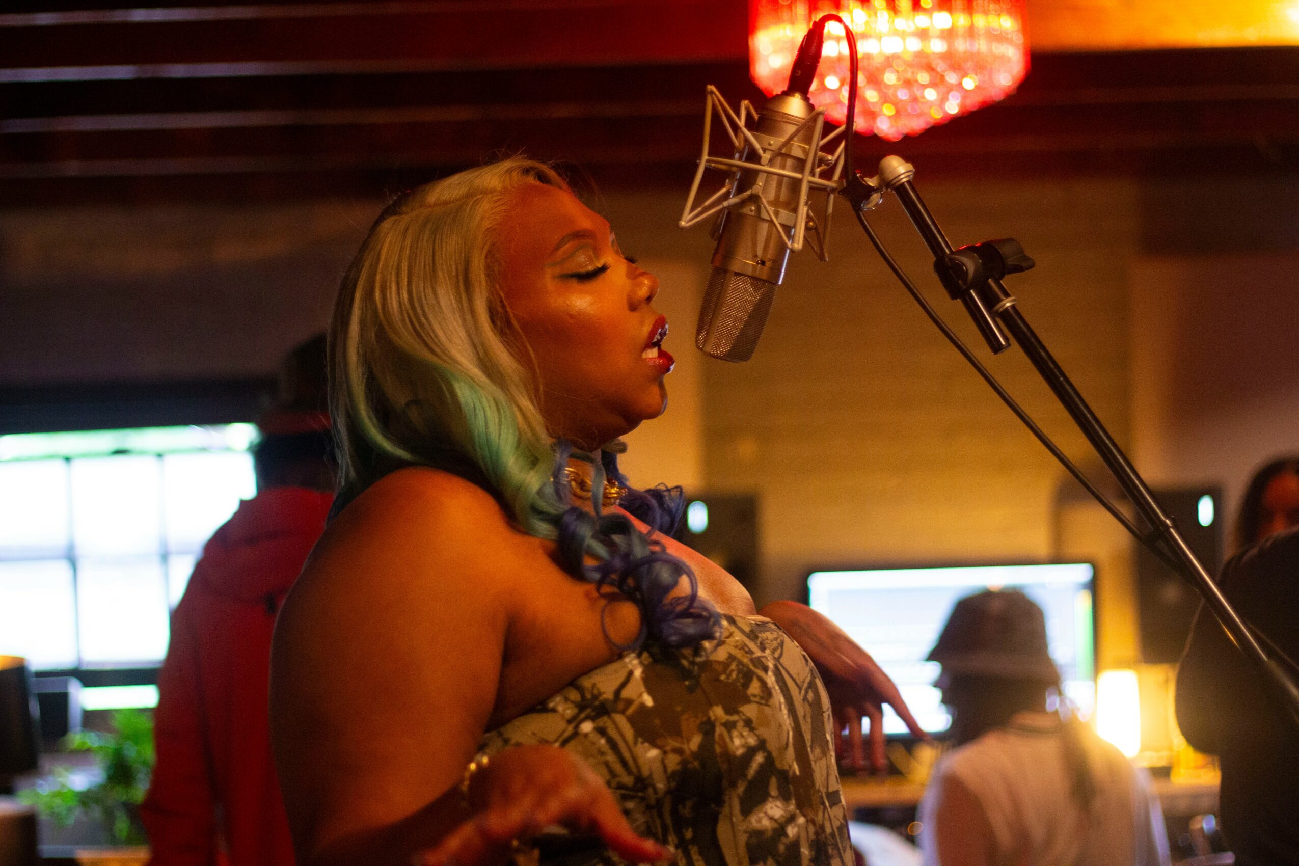 Woman singing into a microphone in a recording studio with warm lighting and people in the background.