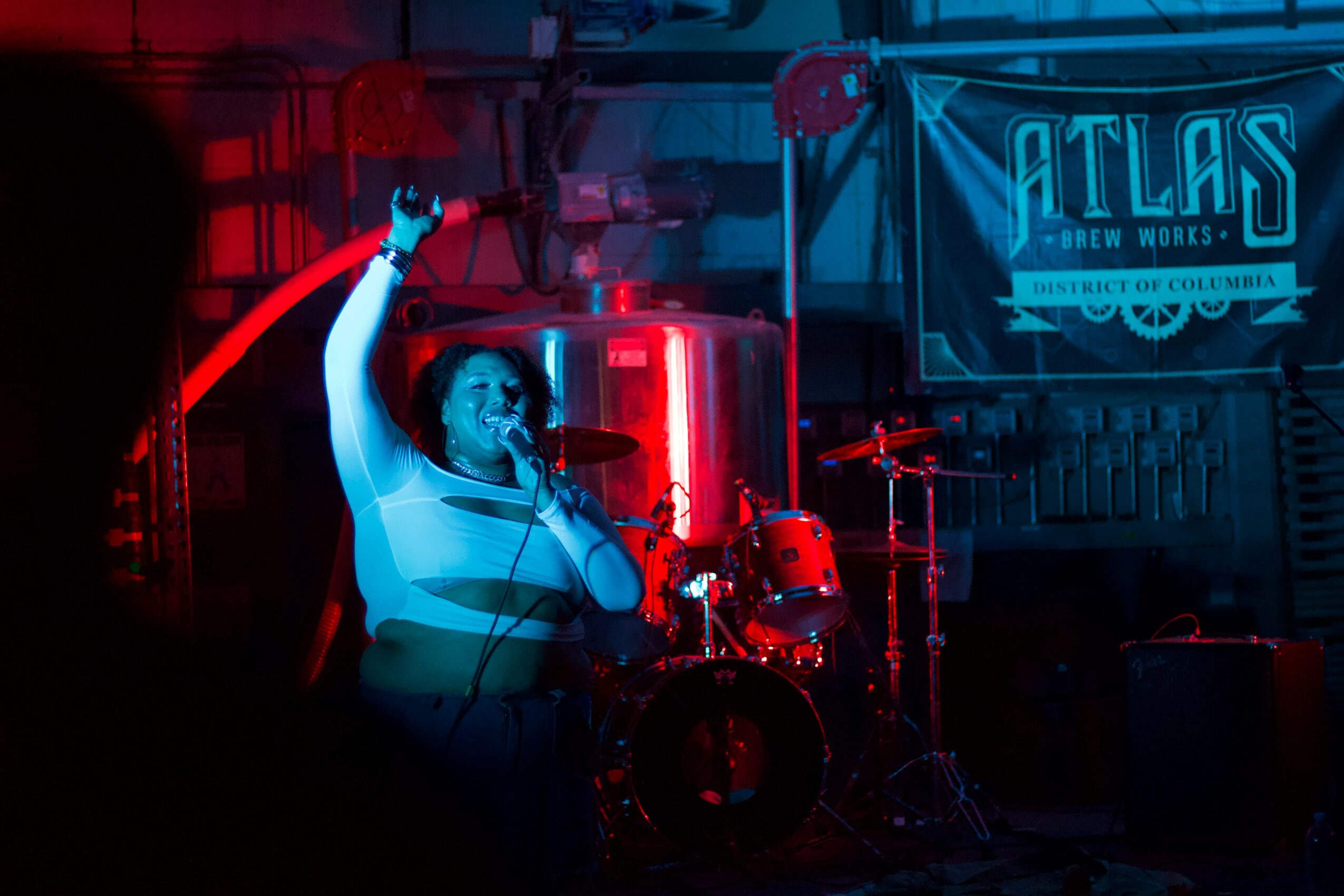 Person singing into microphone with raised hand on stage, drums and a banner in the background, dim lighting.