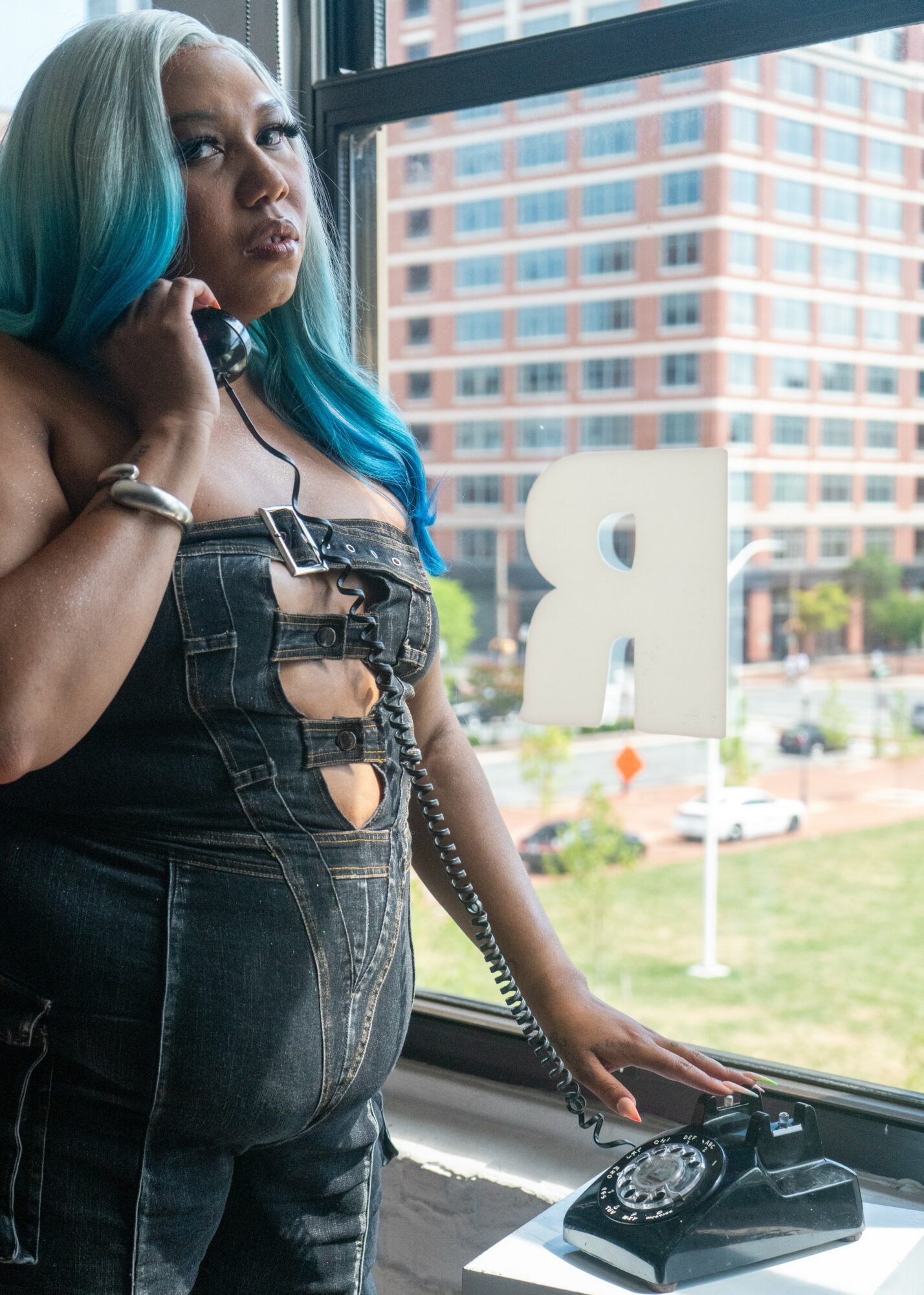 Woman with blue hair talking on a landline phone by a window with city buildings outside.