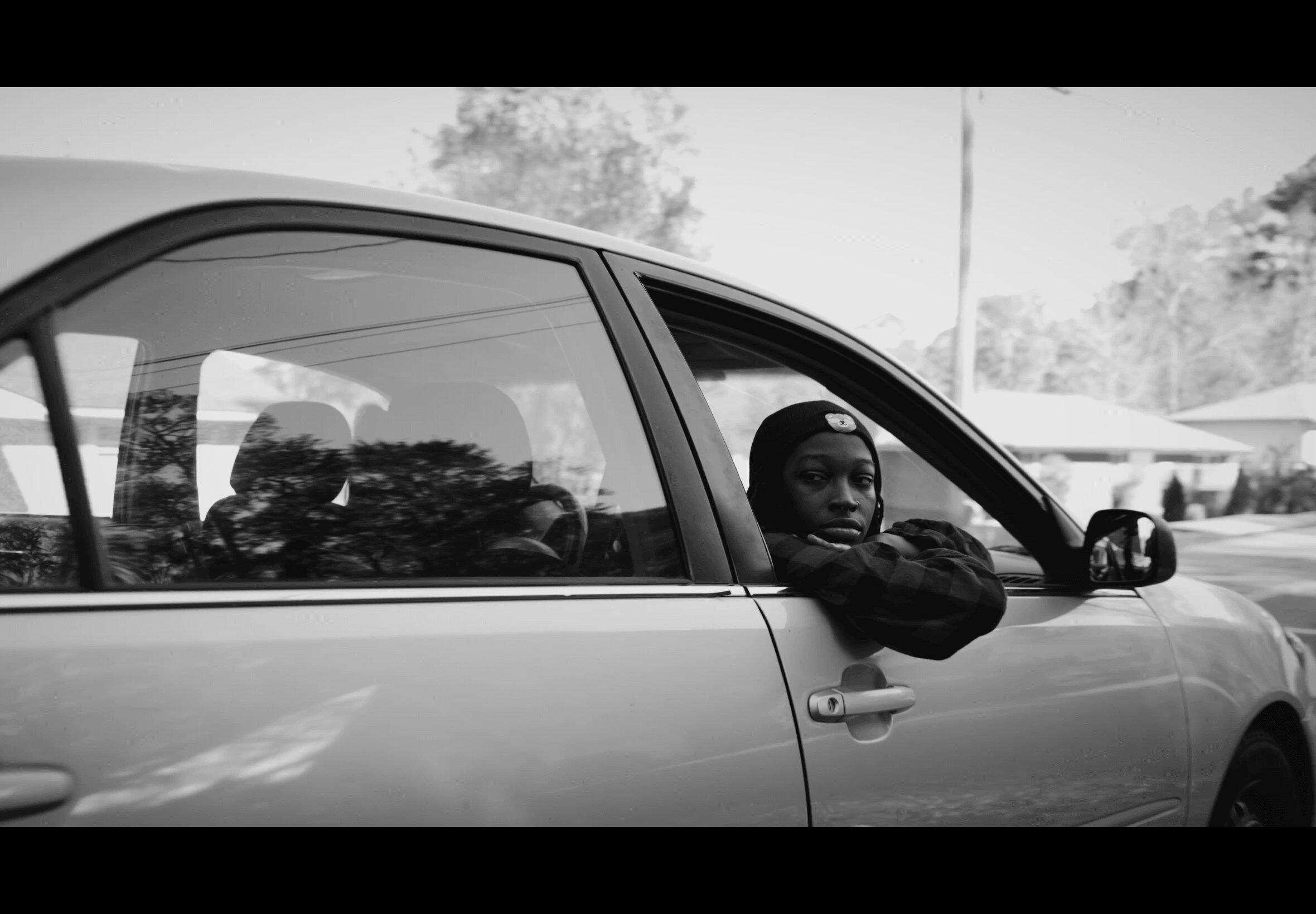 Person looking out of car window with arm resting on window frame, in a parking lot, black and white image.