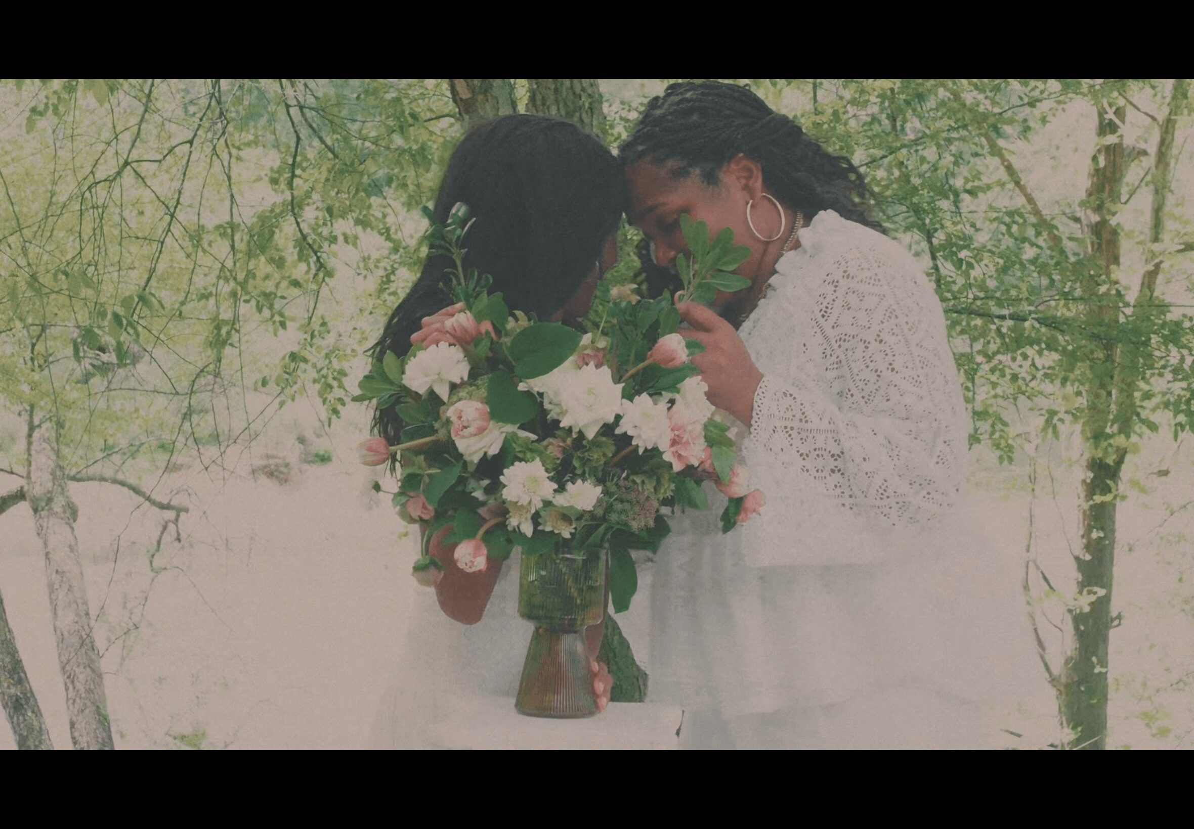 Two women in white dresses hold a large bouquet of flowers outdoors, surrounded by trees and greenery.