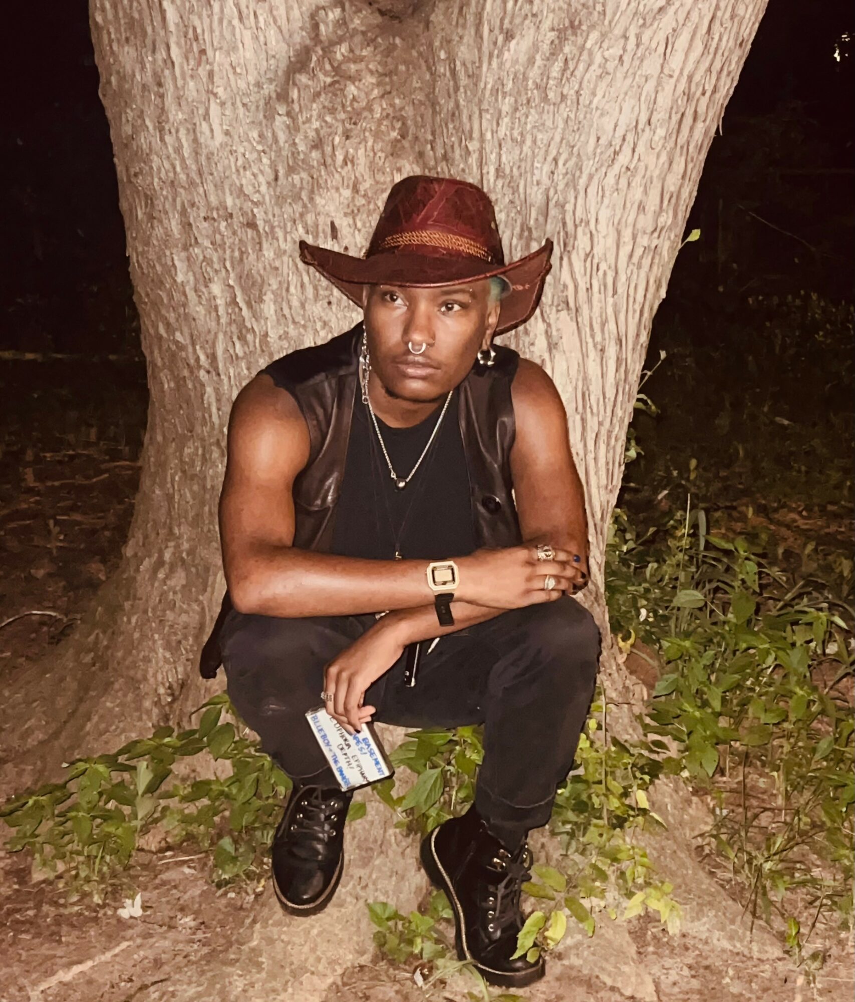 Person crouching outdoors at night, wearing a hat, black sleeveless top, and black pants, with a tree in the background.