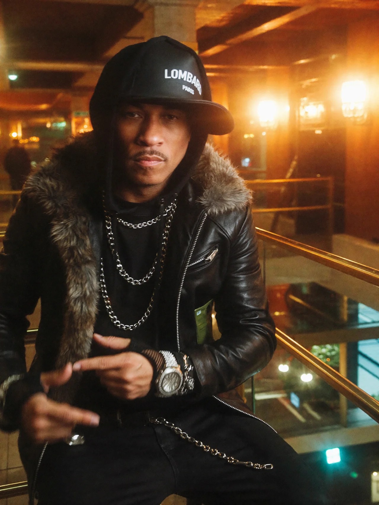 Young man wearing a black cap, leather jacket with fur collar, and jewelry, sitting indoors with warm lighting.