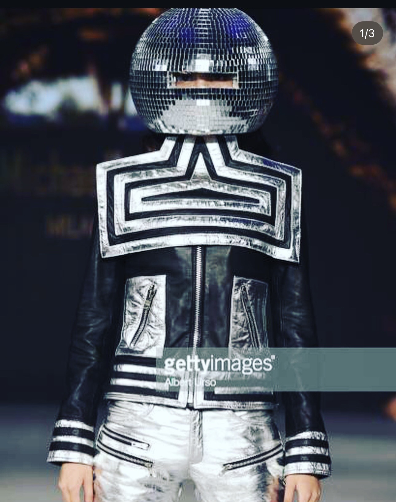 Person wearing a metallic jacket and a disco ball helmet, standing in a dark environment with blurred lights.