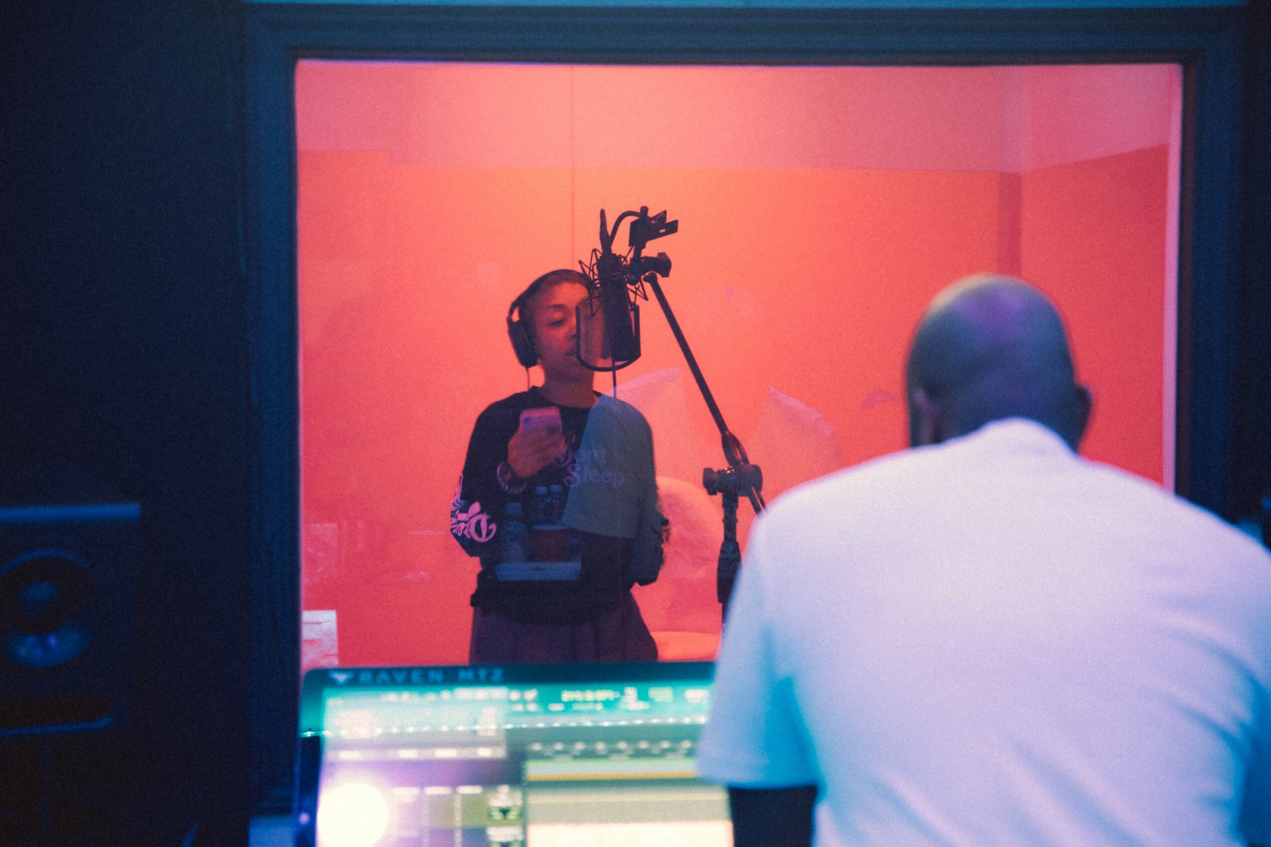 Person in a recording studio singing into a microphone, viewed through a glass window, with a person in a white shirt in foreground.