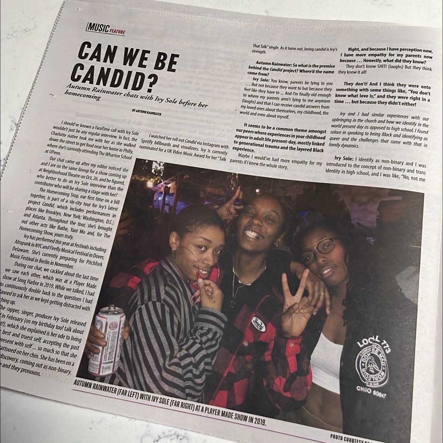 Three people smiling and posing together at a social event, with one making a peace sign gesture. A newspaper article is in the background.