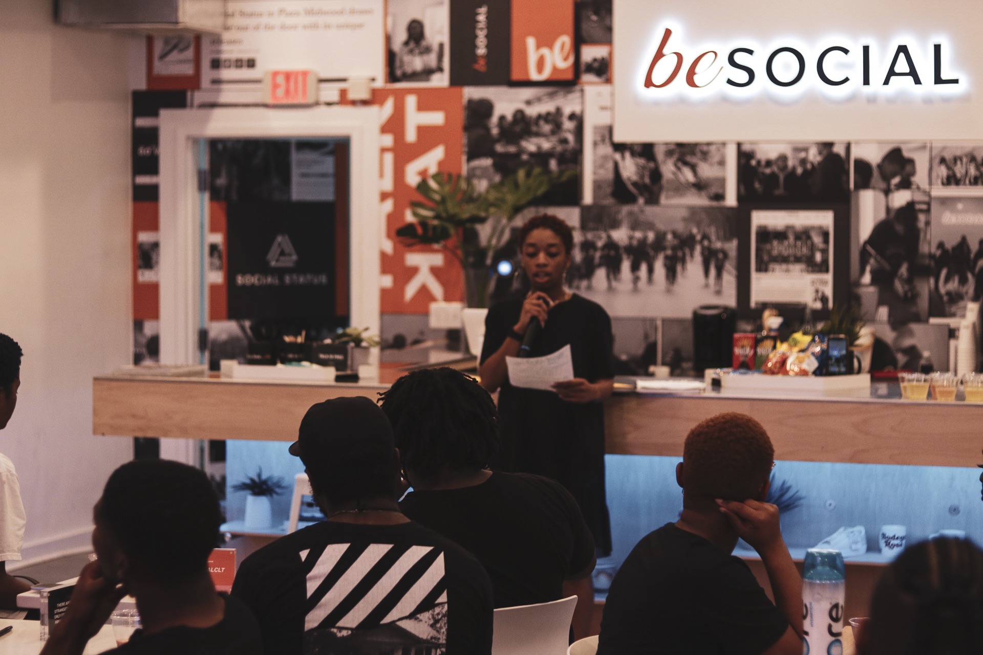 Person speaking at a podium in front of a wall with photos and signs, audience seated facing the speaker.