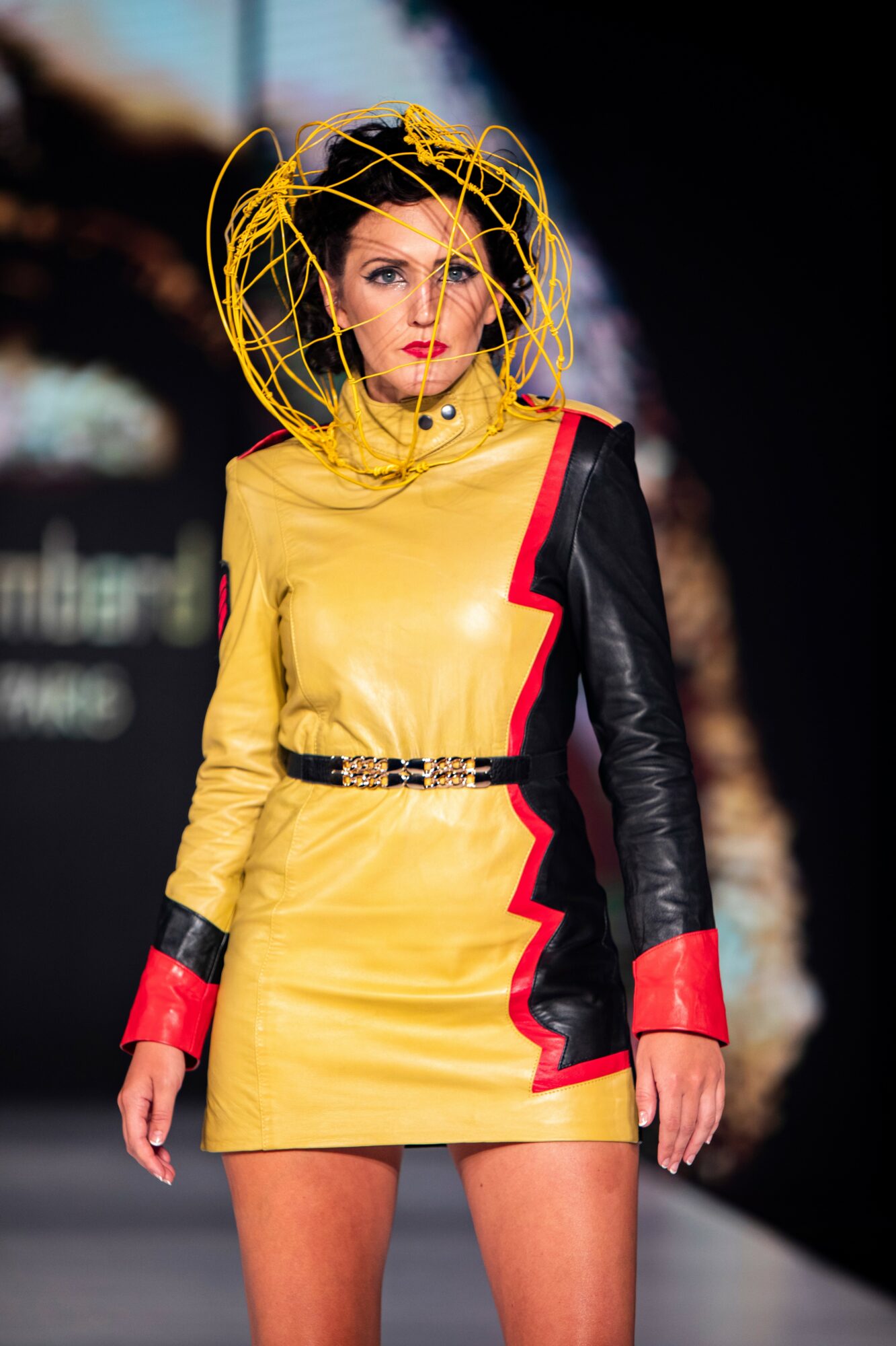 Fashion model walking on runway wearing a yellow dress with black and red accents, and a yellow headpiece.
