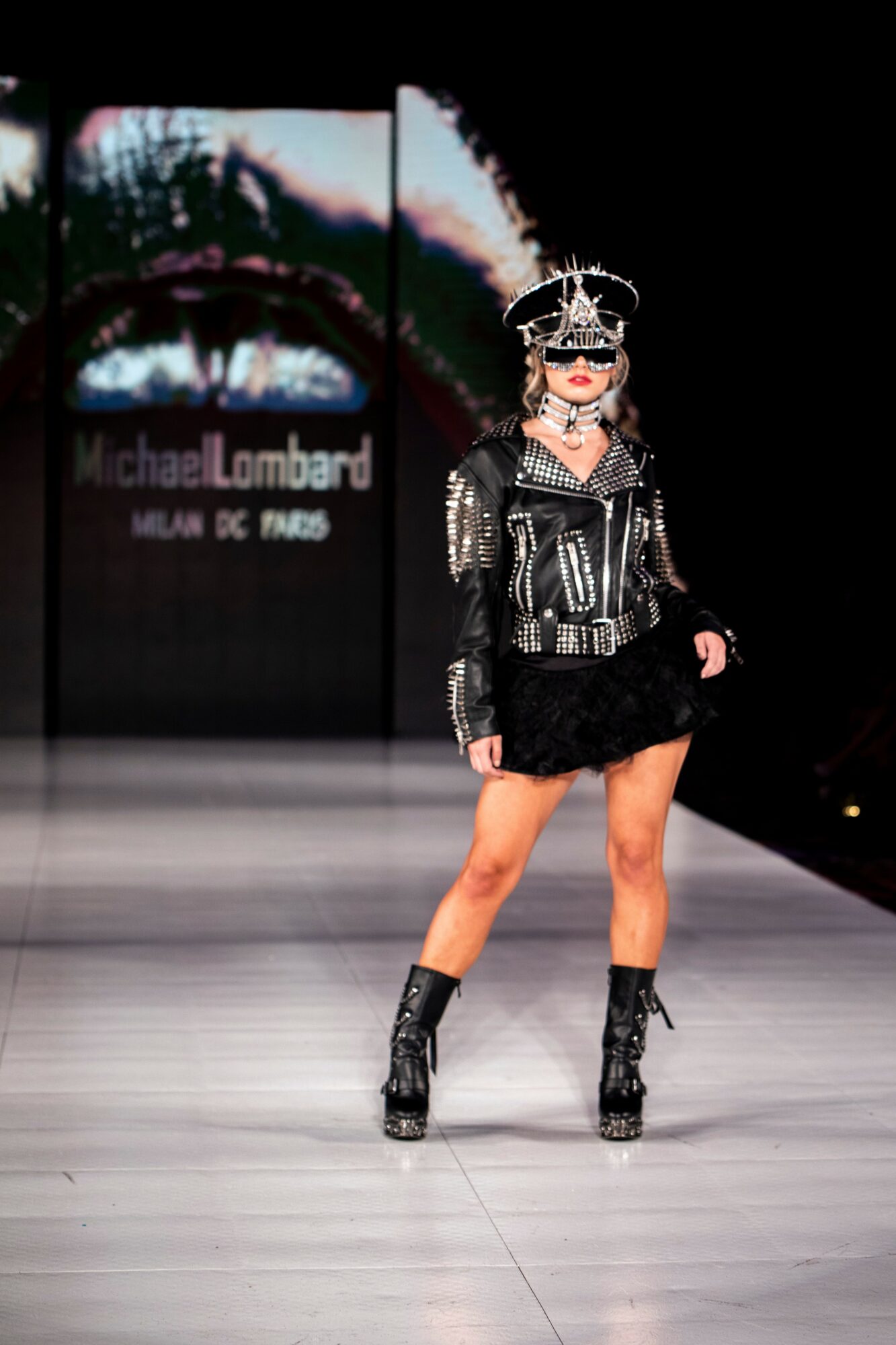 Model walking on runway wearing black leather jacket, black skirt, and high boots, with a large screen displaying text behind her.