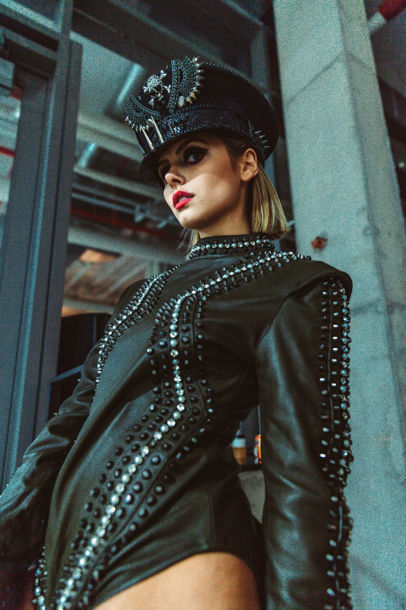 Woman in a black leather outfit with silver embellishments, wearing a matching hat, standing outdoors near a building.