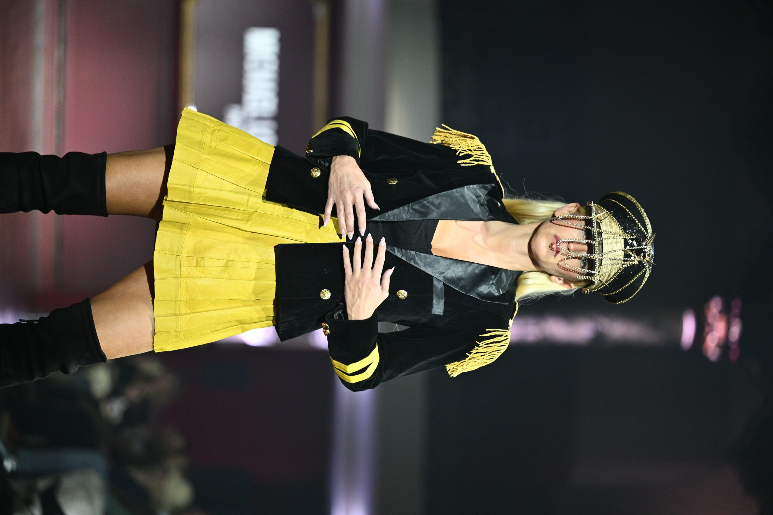 Person in yellow skirt and black jacket with gold accents standing indoors, holding a phone, with blurred background.