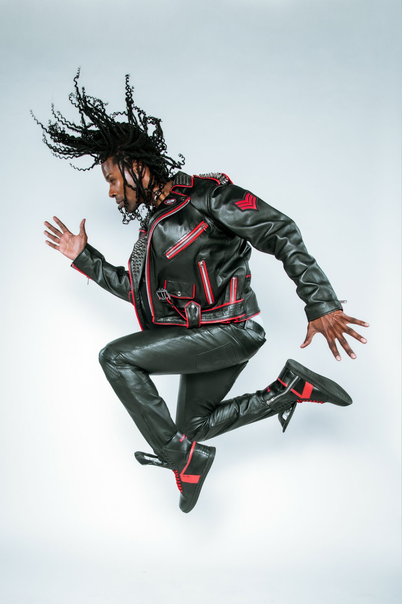 Person jumping mid-air wearing a black leather jacket and pants with red accents, hair flying, against a plain background.