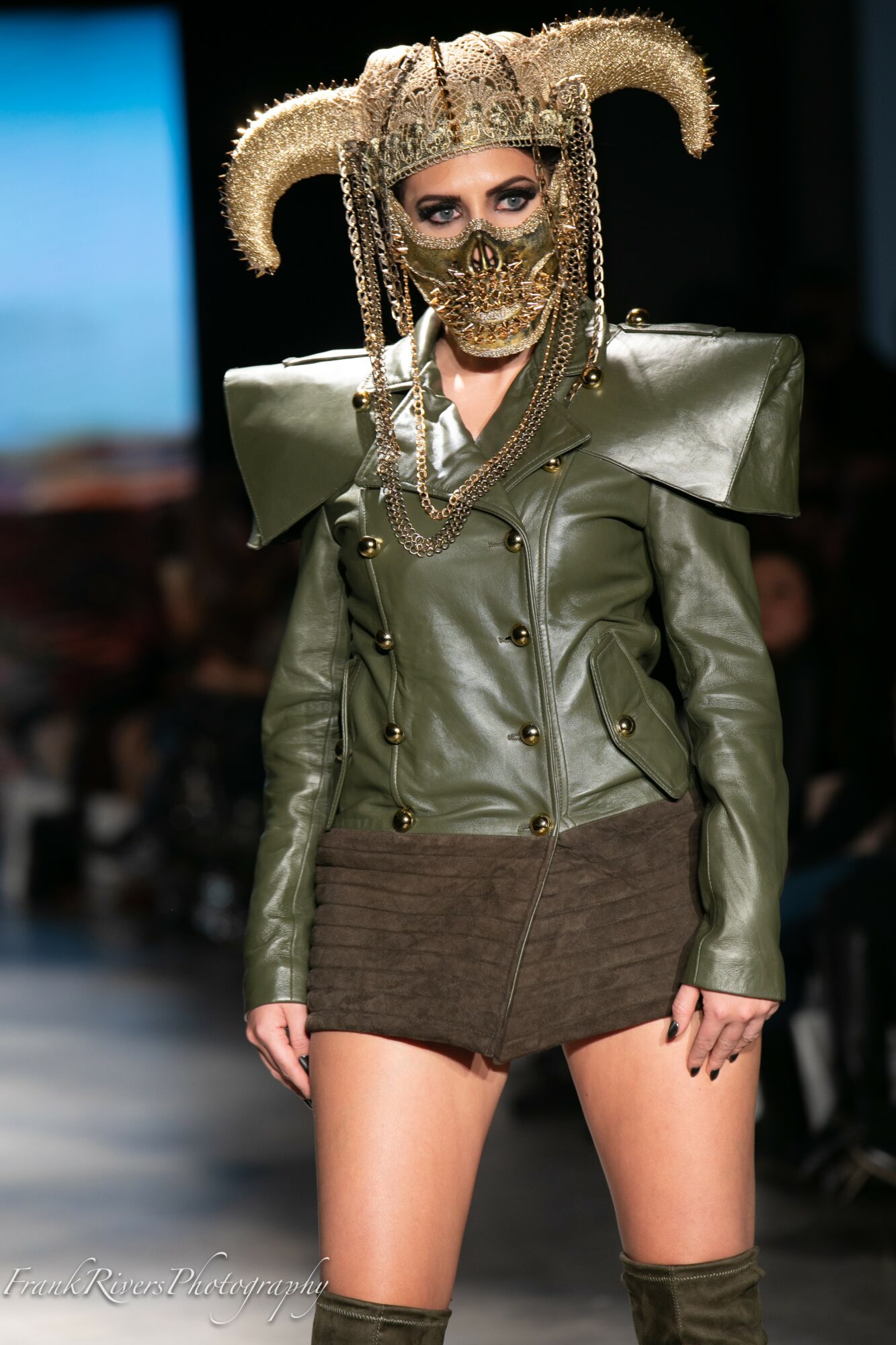 Model walking runway wearing a metallic mask, elaborate headpiece, green leather jacket, and brown skirt.