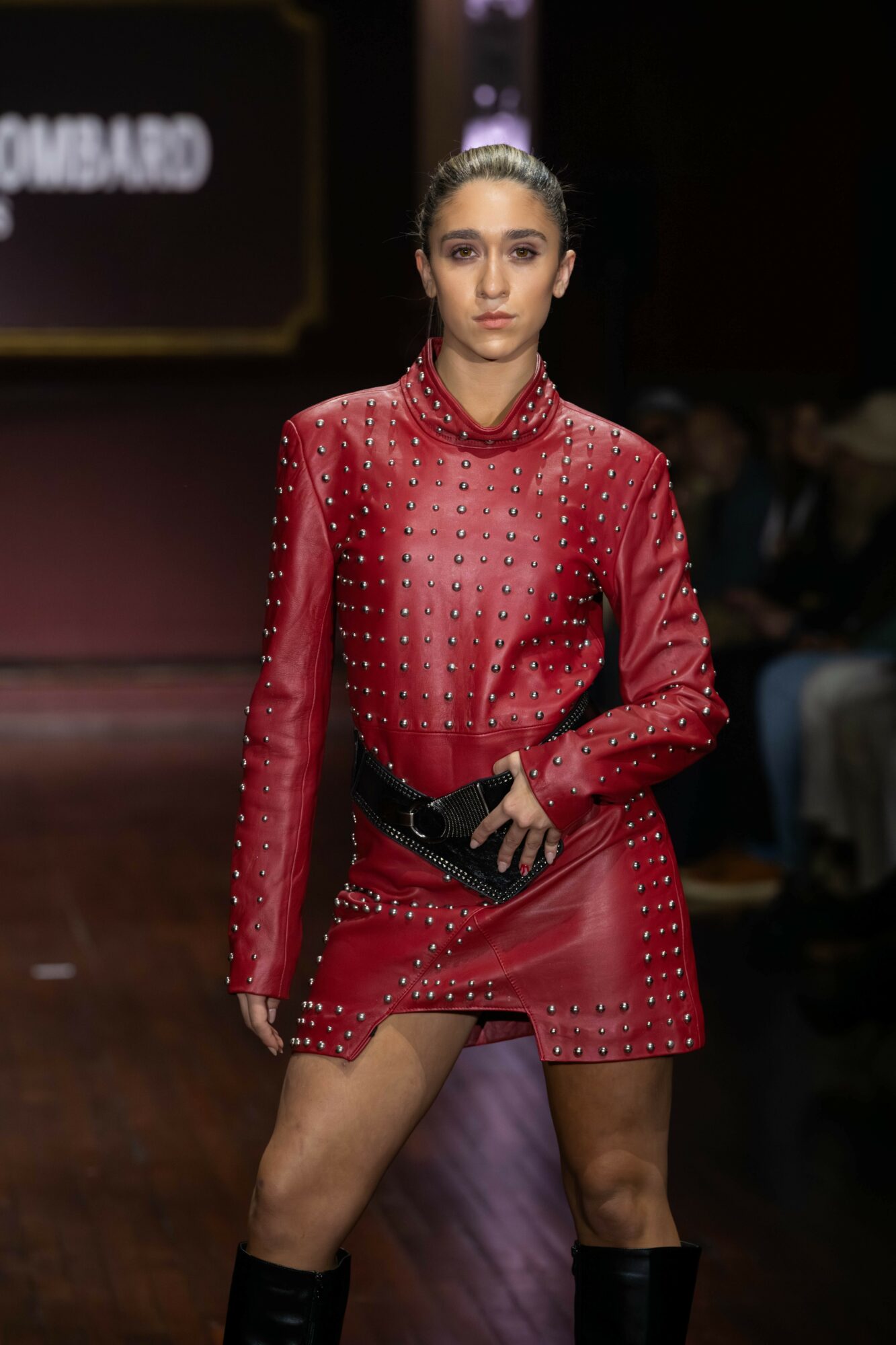 Fashion model walking on runway wearing a red studded leather jacket and skirt, with a black belt and knee-high boots.