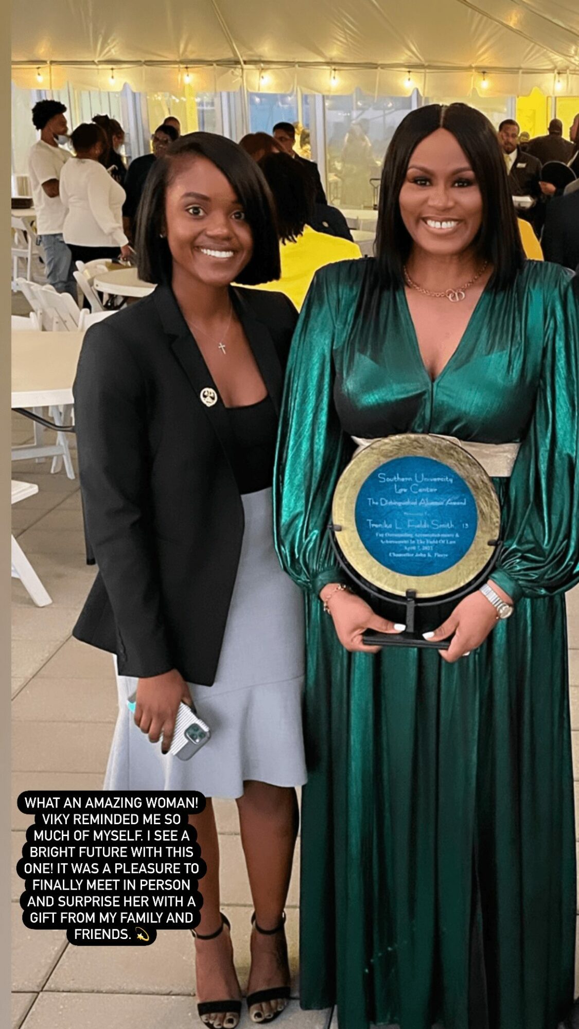 Two women standing indoors, one holding a circular award plaque, smiling, with people in the background at an event.