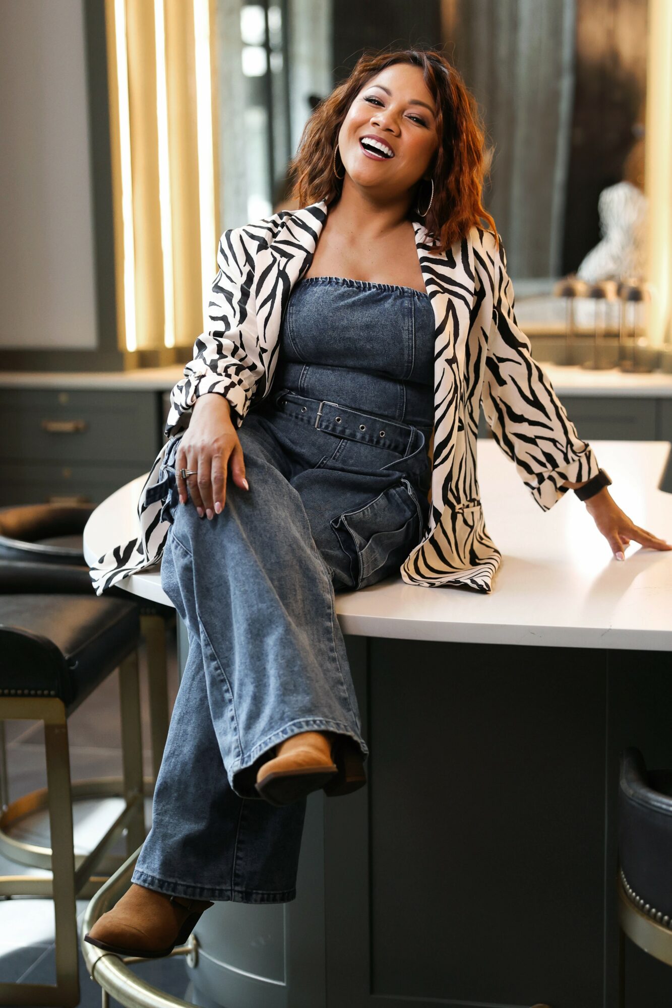 Woman with curly hair smiling, sitting on a kitchen counter, wearing a striped blazer and denim jumpsuit, in a modern kitchen.