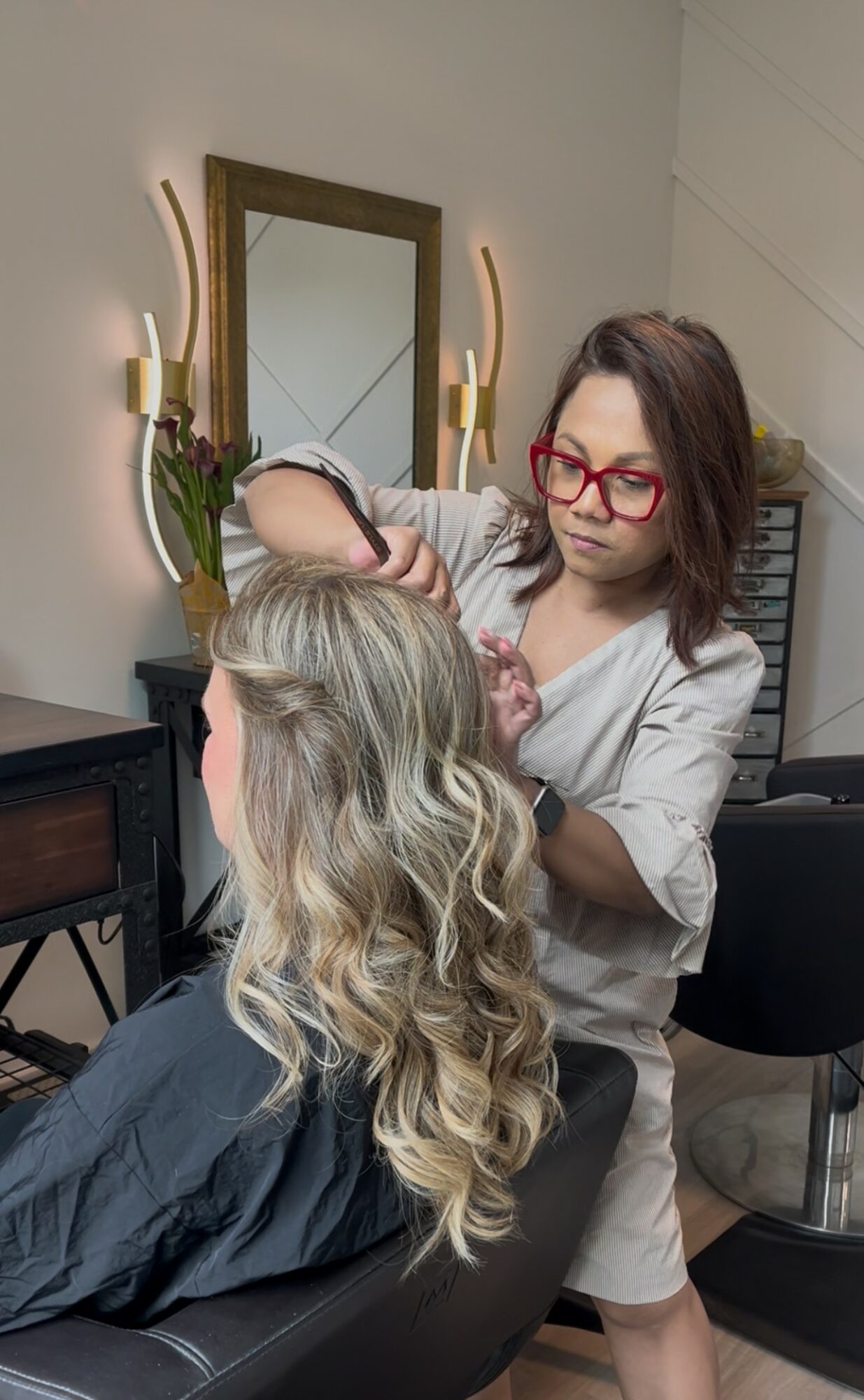 Hairdresser with red glasses styling a client's long, wavy blonde hair in a salon. Mirror and lighting visible in background.