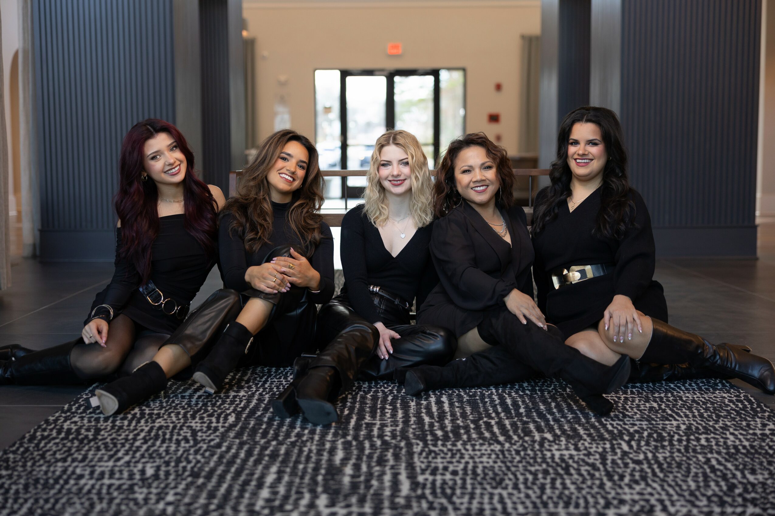 Five women sitting on the floor in a modern indoor space, smiling and posing for the photo.