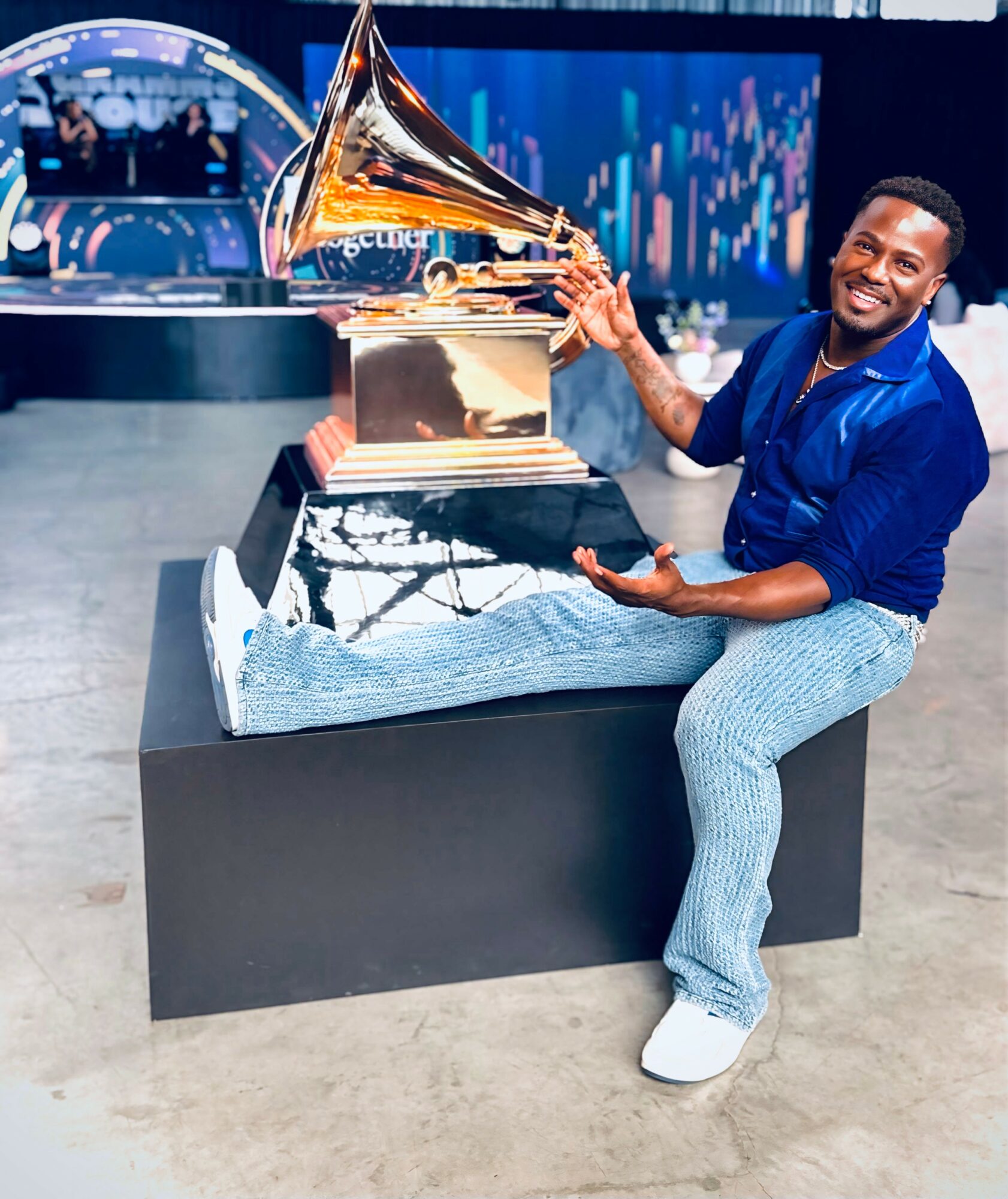 Man smiling with a Grammy award trophy, sitting on a black platform, in a modern indoor setting.