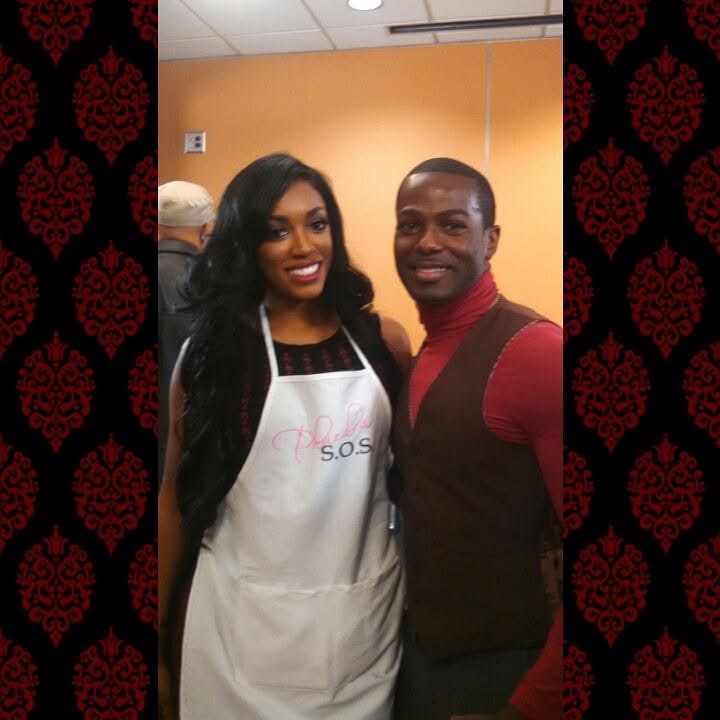 Two people smiling, one wearing an apron, standing indoors against a yellow wall.