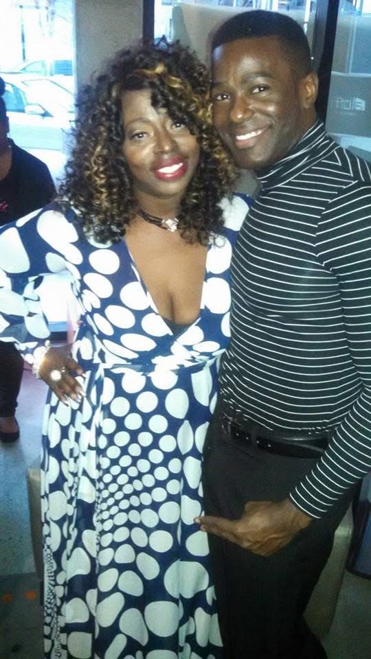 Two people smiling, standing close together indoors, woman in a blue and white polka dot dress, man in a striped shirt.