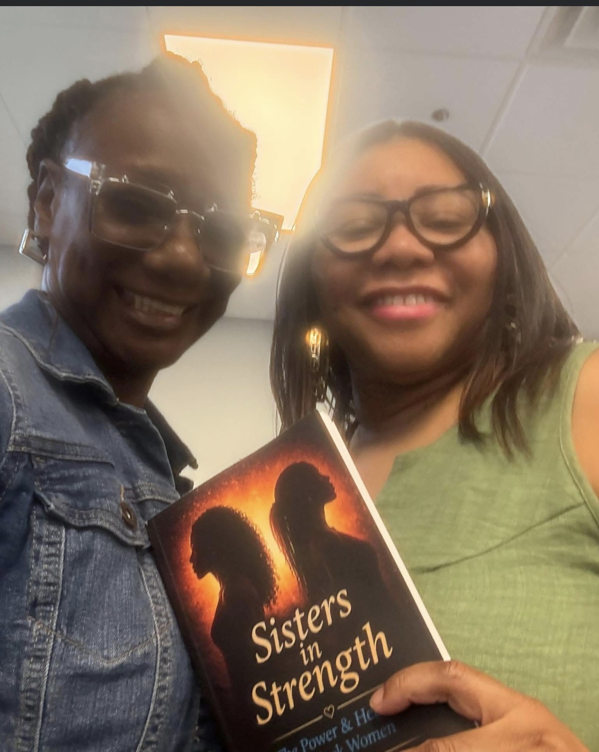 Two women smiling, holding a book titled 'Sisters' Strength' with silhouettes of two women on the cover.