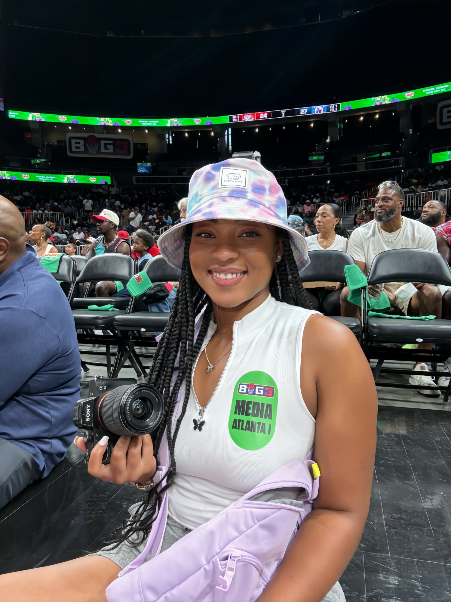 Smiling woman with braided hair wearing a bucket hat and media badge, holding a camera at an indoor event.
