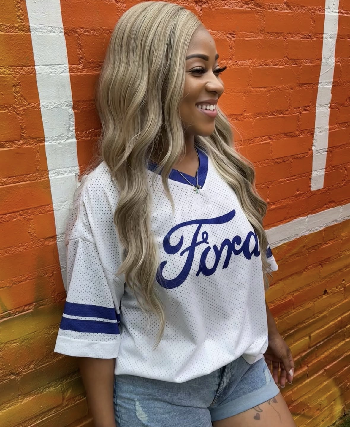Smiling woman with long wavy blonde hair wearing a white Ford jersey and denim shorts, standing against an orange brick wall.