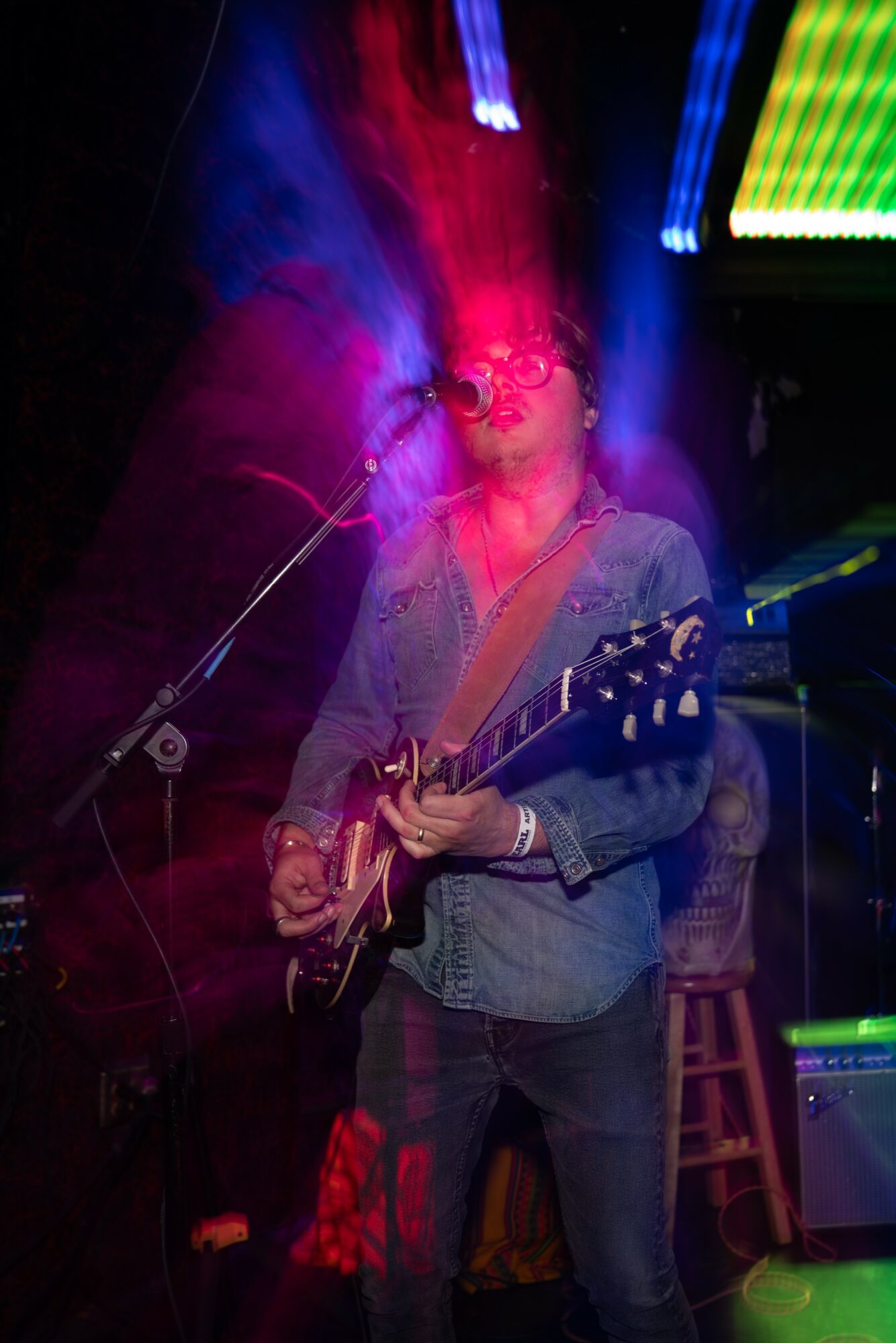 Person playing guitar on stage with colorful lights and smoke, wearing glasses and a denim jacket.