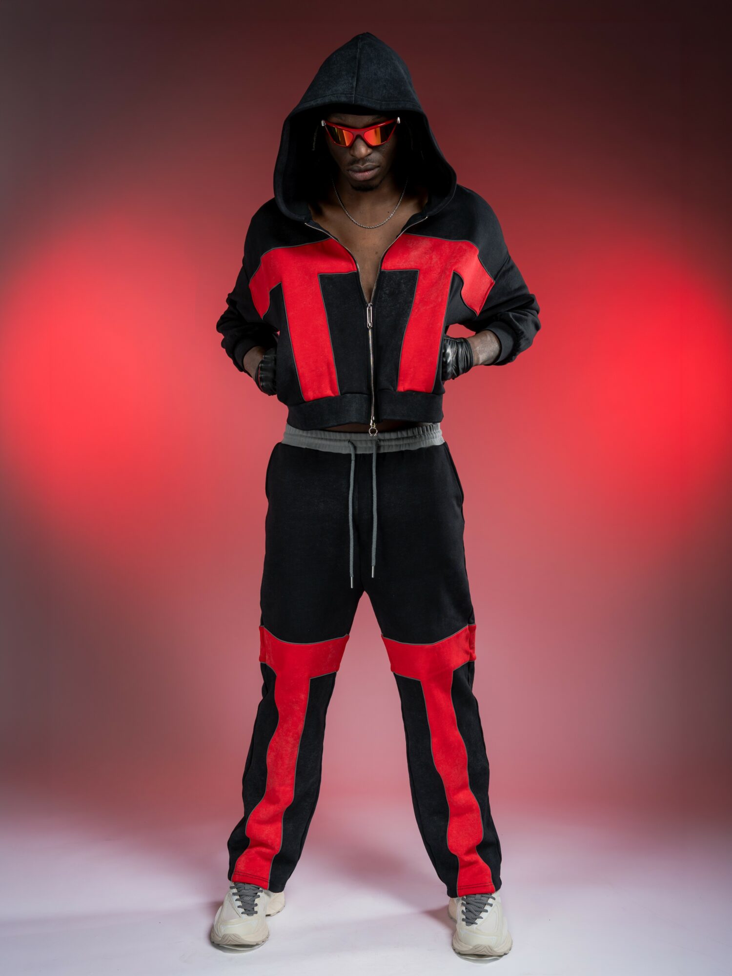 Person wearing a black and red tracksuit with a hood, sunglasses, and sneakers, standing against a red gradient background.