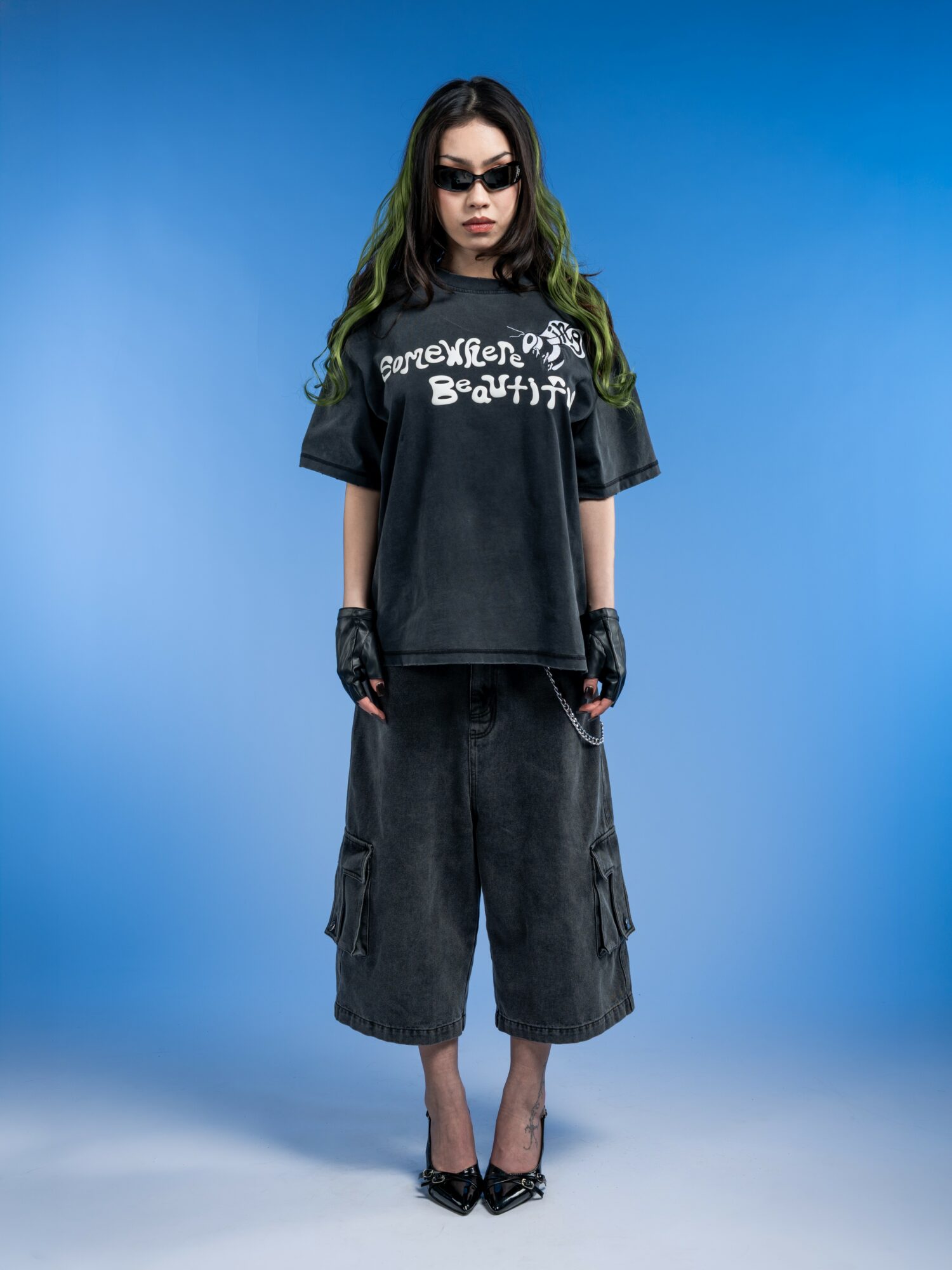 Young woman with long dark hair, sunglasses, black oversized t-shirt, baggy shorts, standing against blue background.