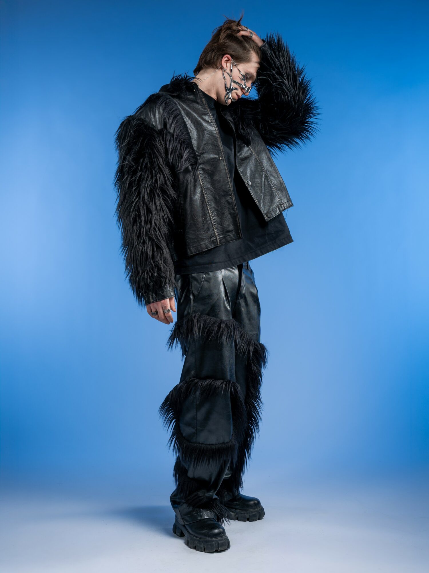 Person in black furry jacket and pants, standing sideways with head tilted down, against a blue background.