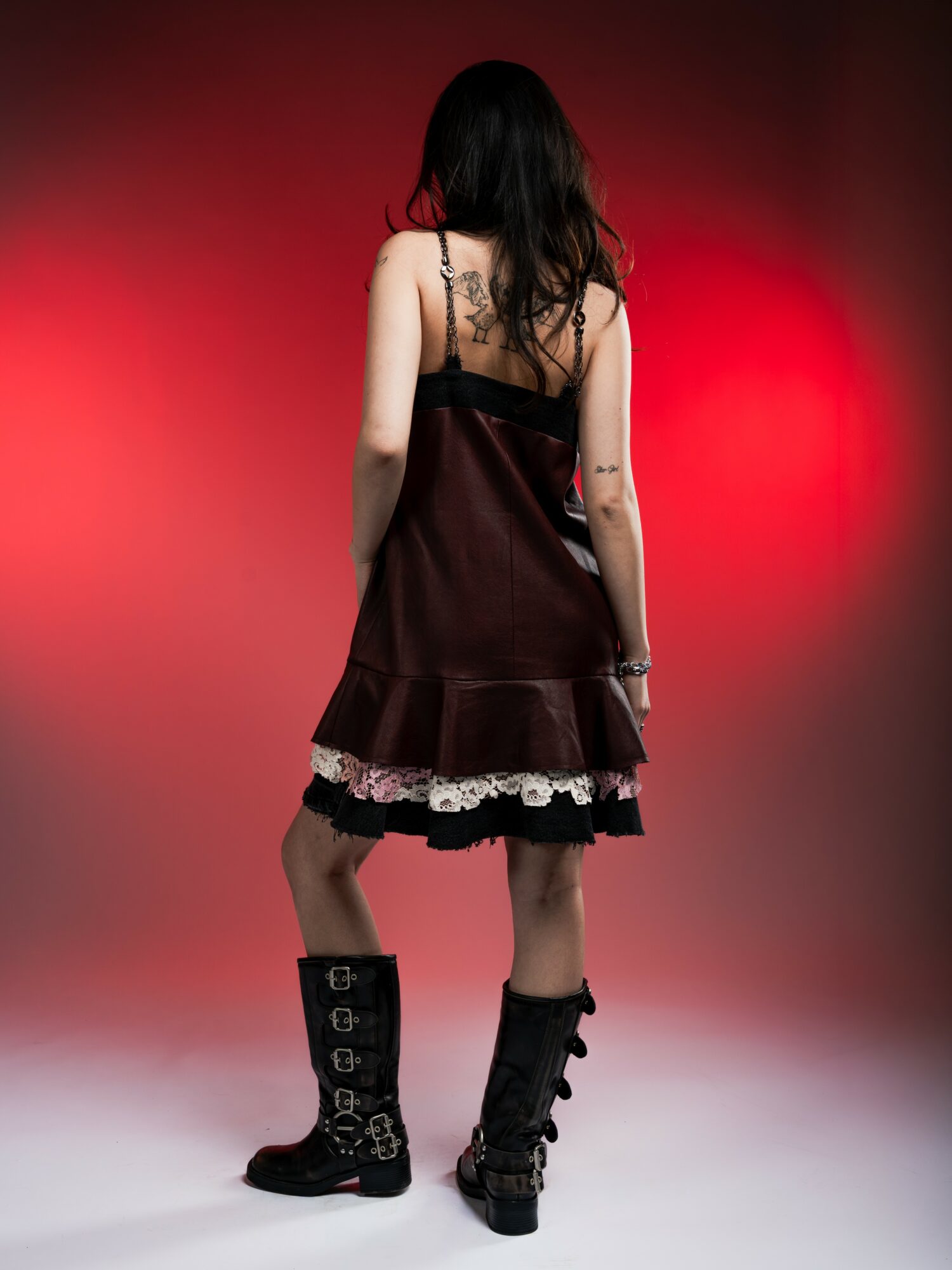Person with long dark hair wearing a dark dress with lace trim, black boots, standing against a red gradient background.