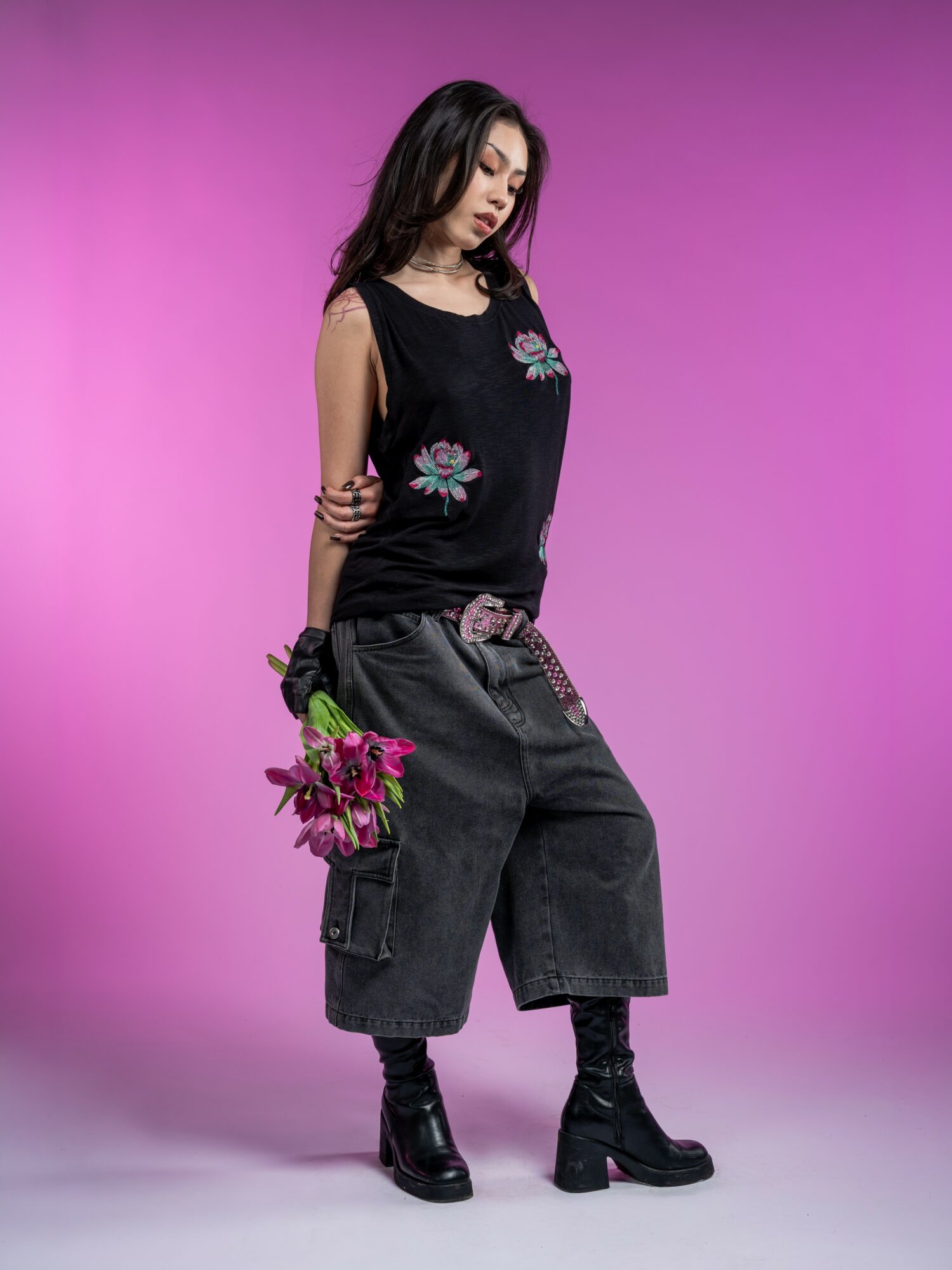 Young woman with dark hair in a black sleeveless top and wide black pants holding pink flowers against a pink background.