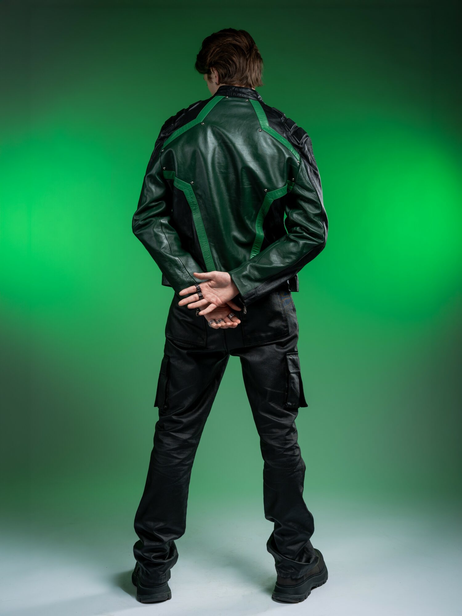 Person standing with back facing camera, hands behind back, wearing a black leather jacket and pants, green background.