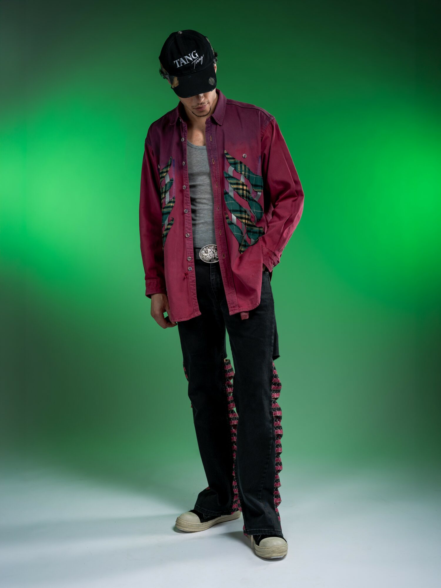 Young man wearing a black cap, maroon jacket, black pants, and sneakers, standing against a green background.