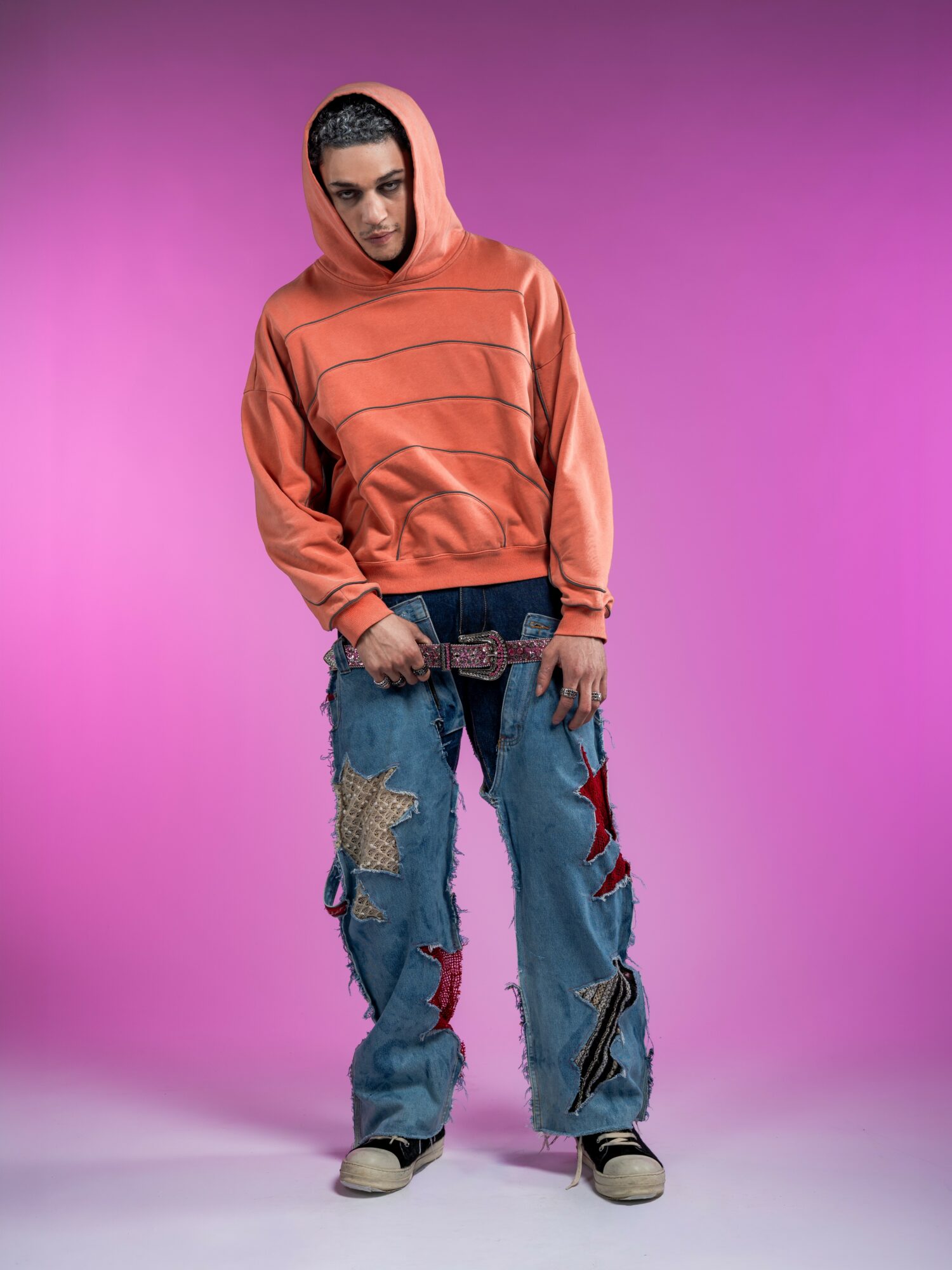 Person wearing an orange hoodie and baggy jeans with patches, standing against a pink background.
