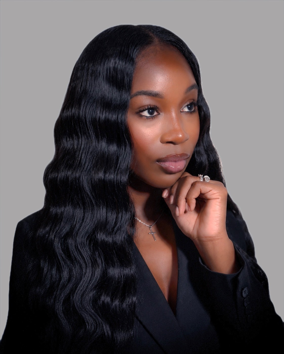 Portrait of a woman with long, wavy black hair, wearing a black blazer, resting her chin on her hand, against a gray background.