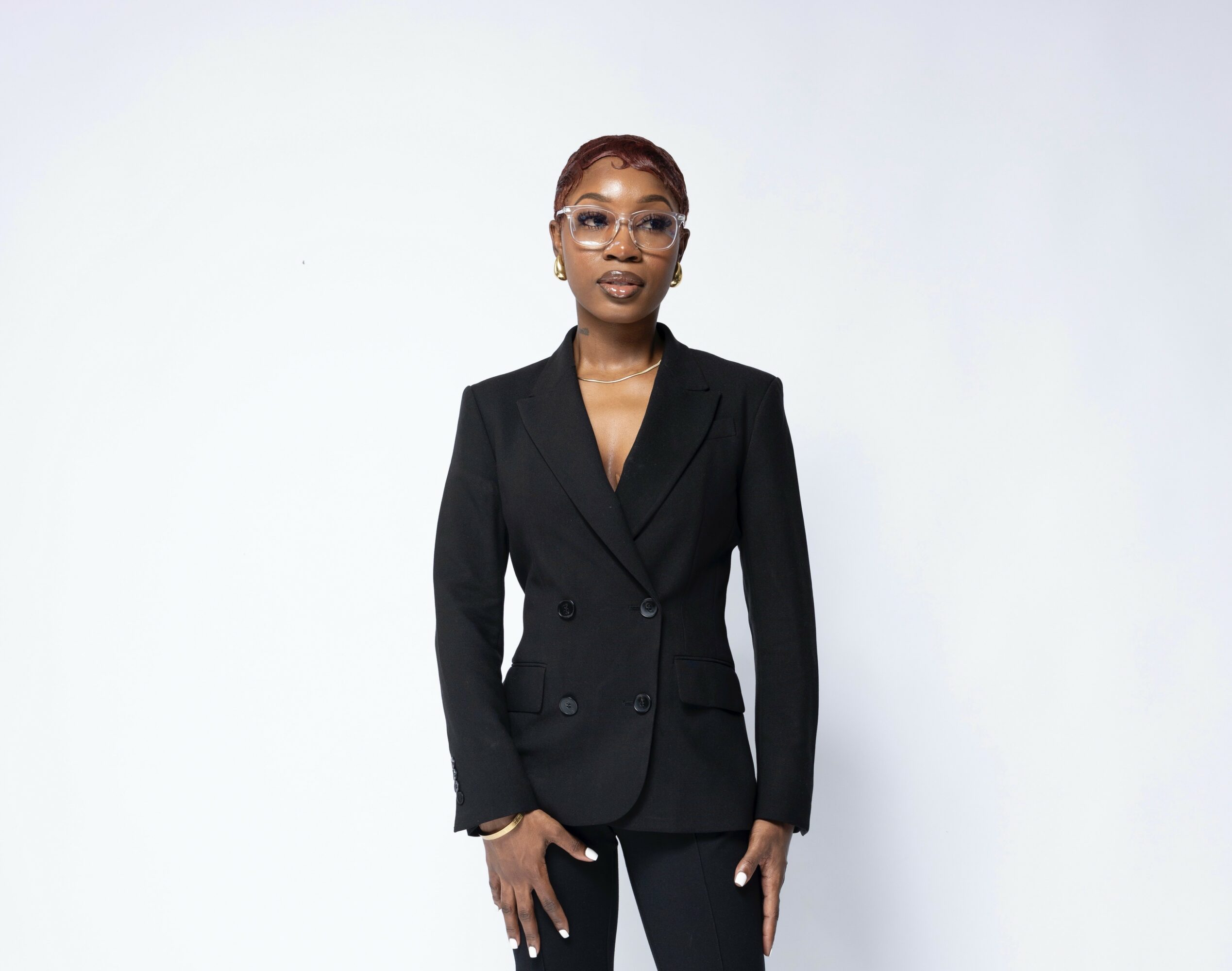 A woman in a black blazer and glasses stands against a plain light background.