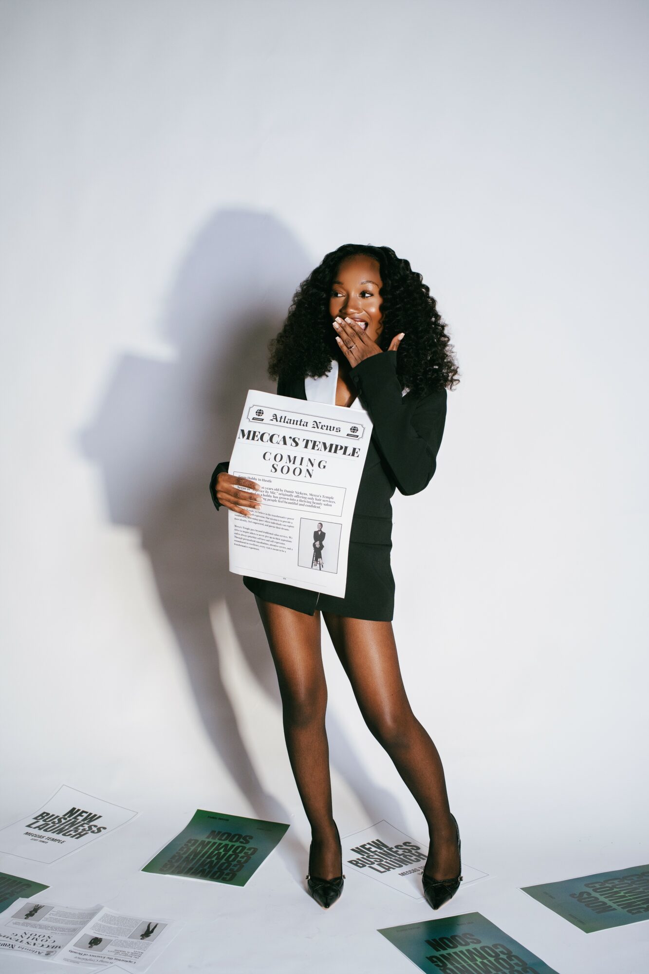 Woman in black dress and tights holding a newspaper, covering her mouth with her hand, standing against a plain white background.