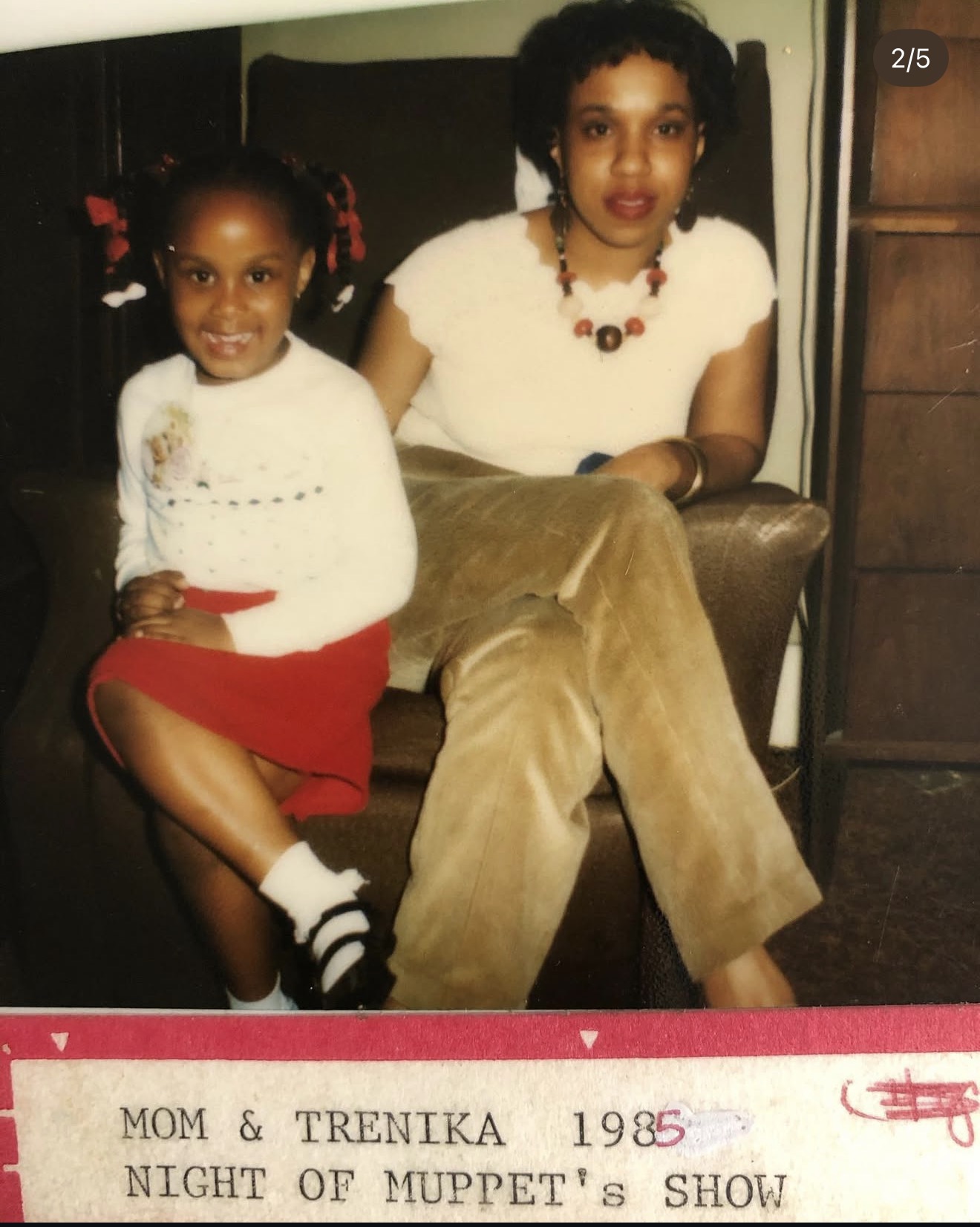 Two girls sitting on a chair, one young girl smiling with pigtails and a red skirt, and an older girl with curly hair and a necklace, sitting with crossed legs.