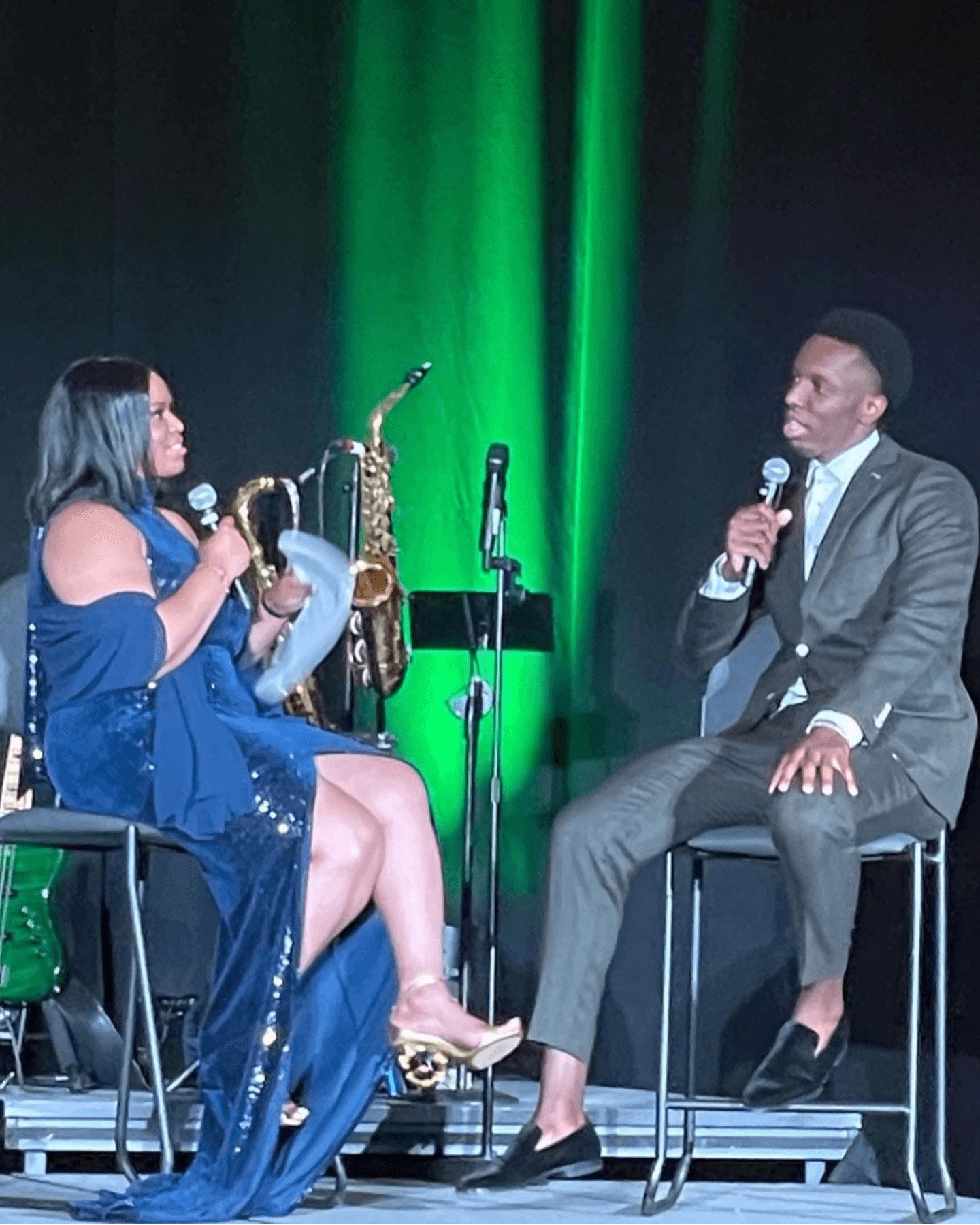 Two people sit on stage, one holding a saxophone and a paper, the other speaking into a microphone, with green stage lighting.
