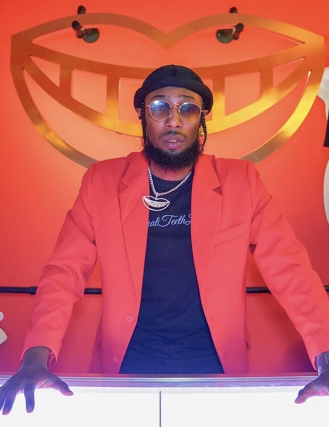 Man with glasses and a black cap wearing a red blazer, standing in front of a large smiling face logo on a red background.