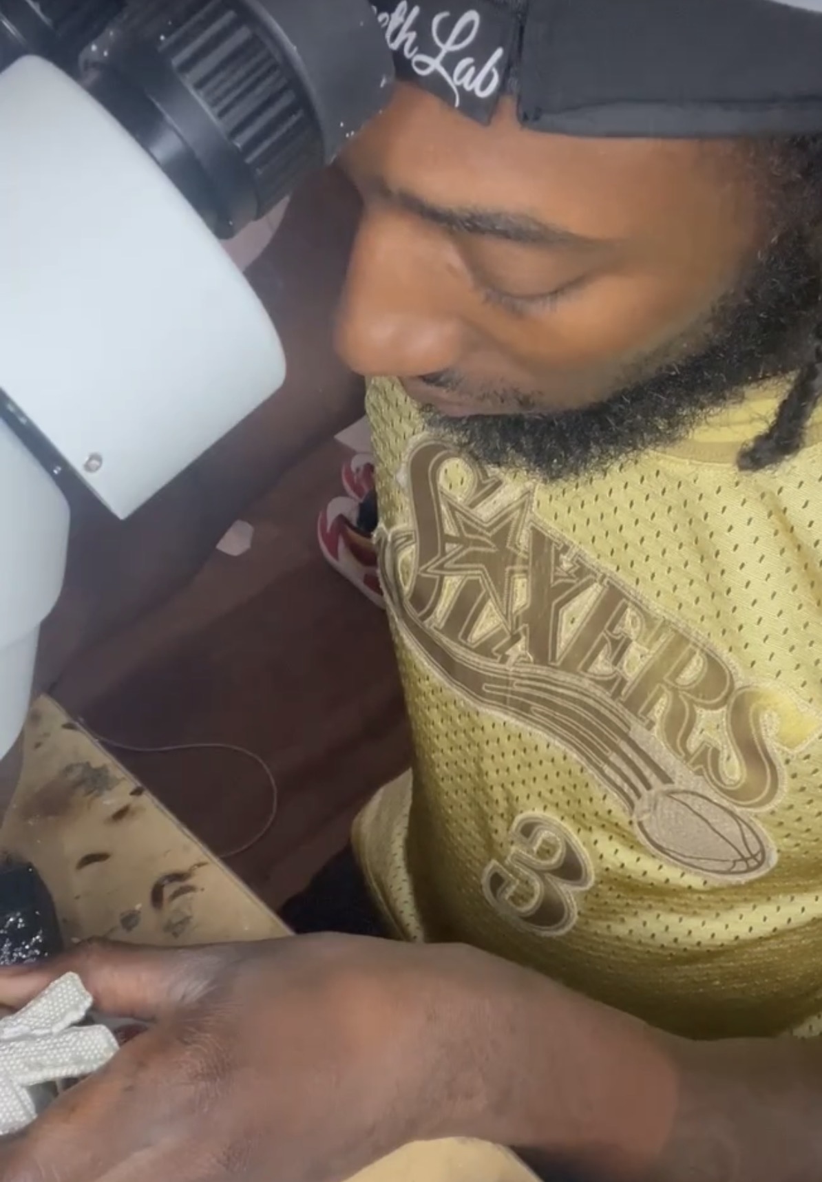 Person looking into a microscope, wearing a yellow shirt and a cap, with hand on a surface.