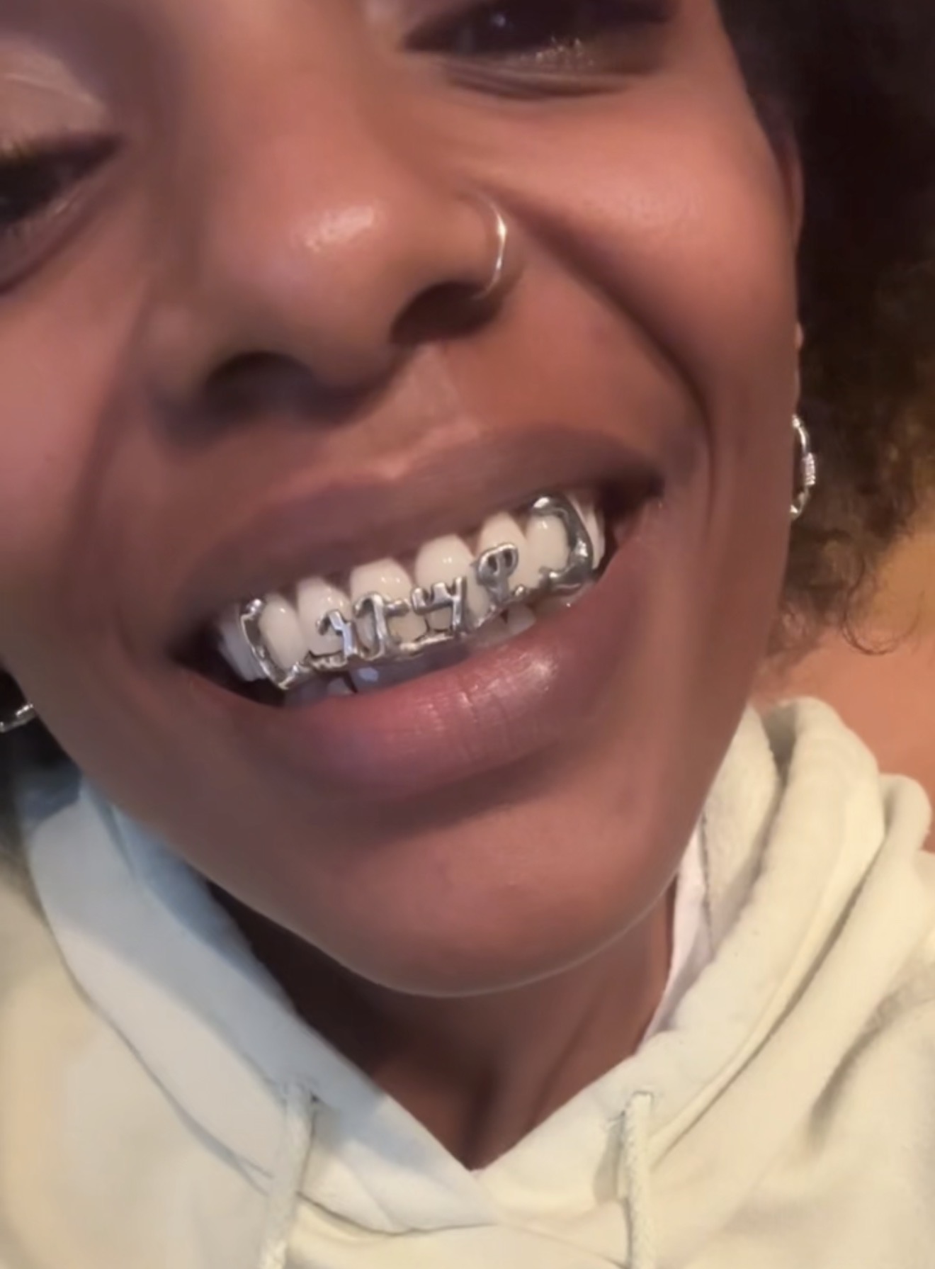 Close-up of a smiling person with a nose ring and a grill on their teeth, wearing a light-colored hoodie.