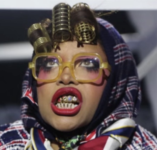 Person with curlers in hair, wearing large yellow glasses, red lipstick, and a patterned headscarf, showing teeth with grillz.