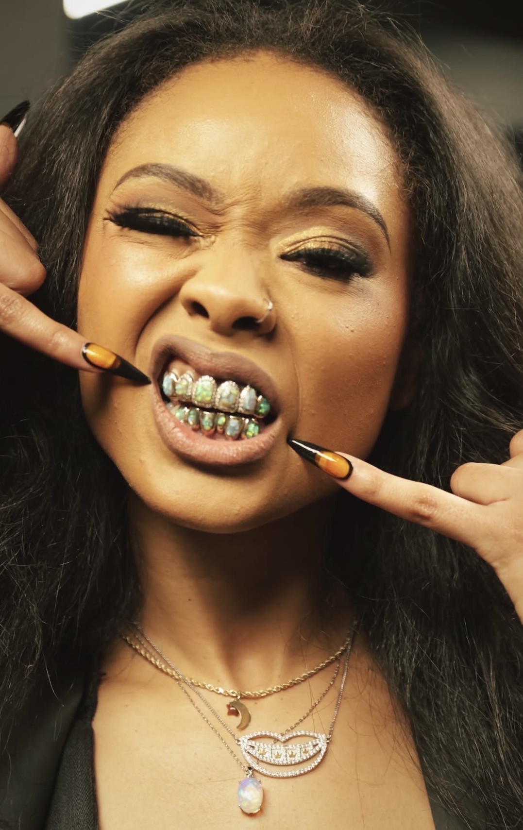Woman with dark hair showing teeth with grills, pointing at her mouth with both hands, making a playful expression.