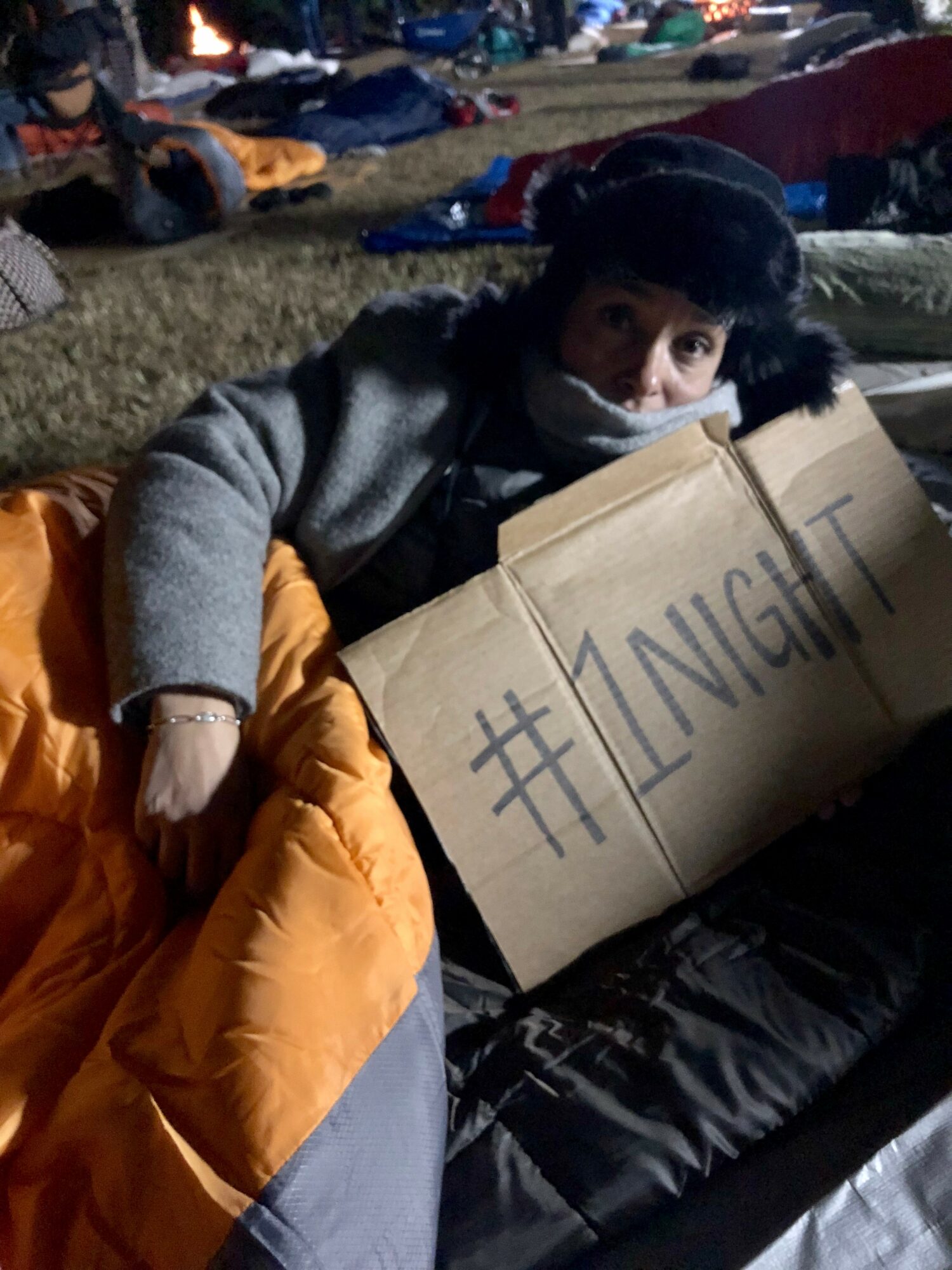 Child holding a cardboard sign with '#1NIGHT' written on it, sitting outdoors at night with other people in the background.